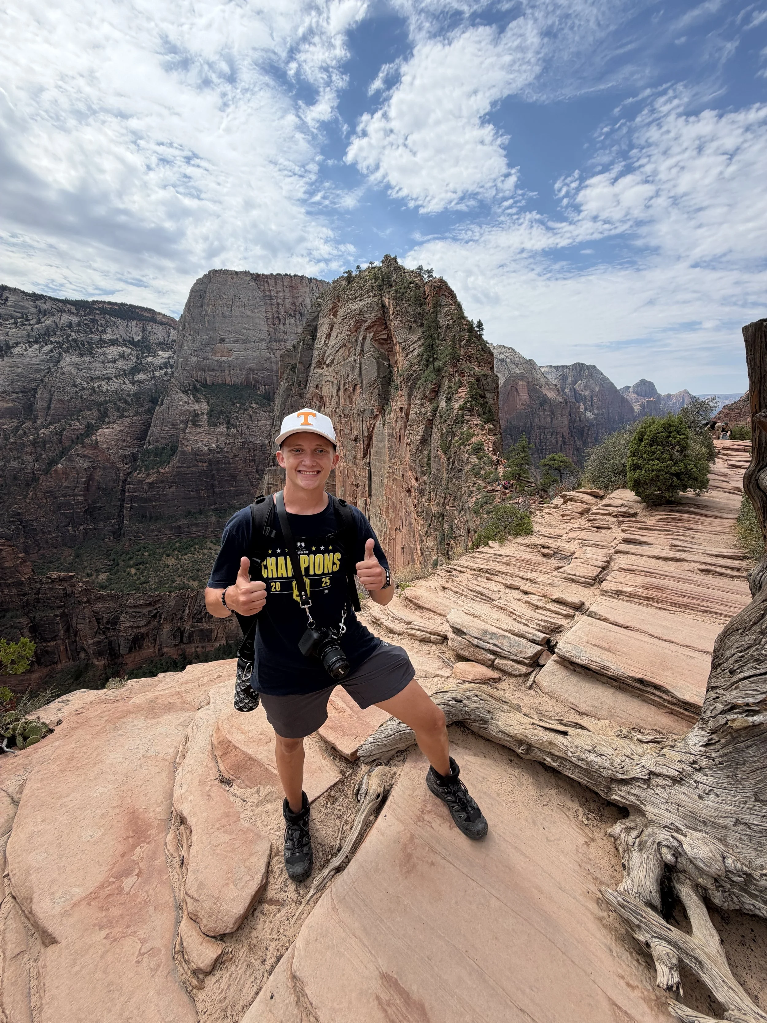 Young man with a backpack and a camera smiling and giving a thumbs-up at Zion National Park, with tall red rock formations and a partly cloudy sky in the background.