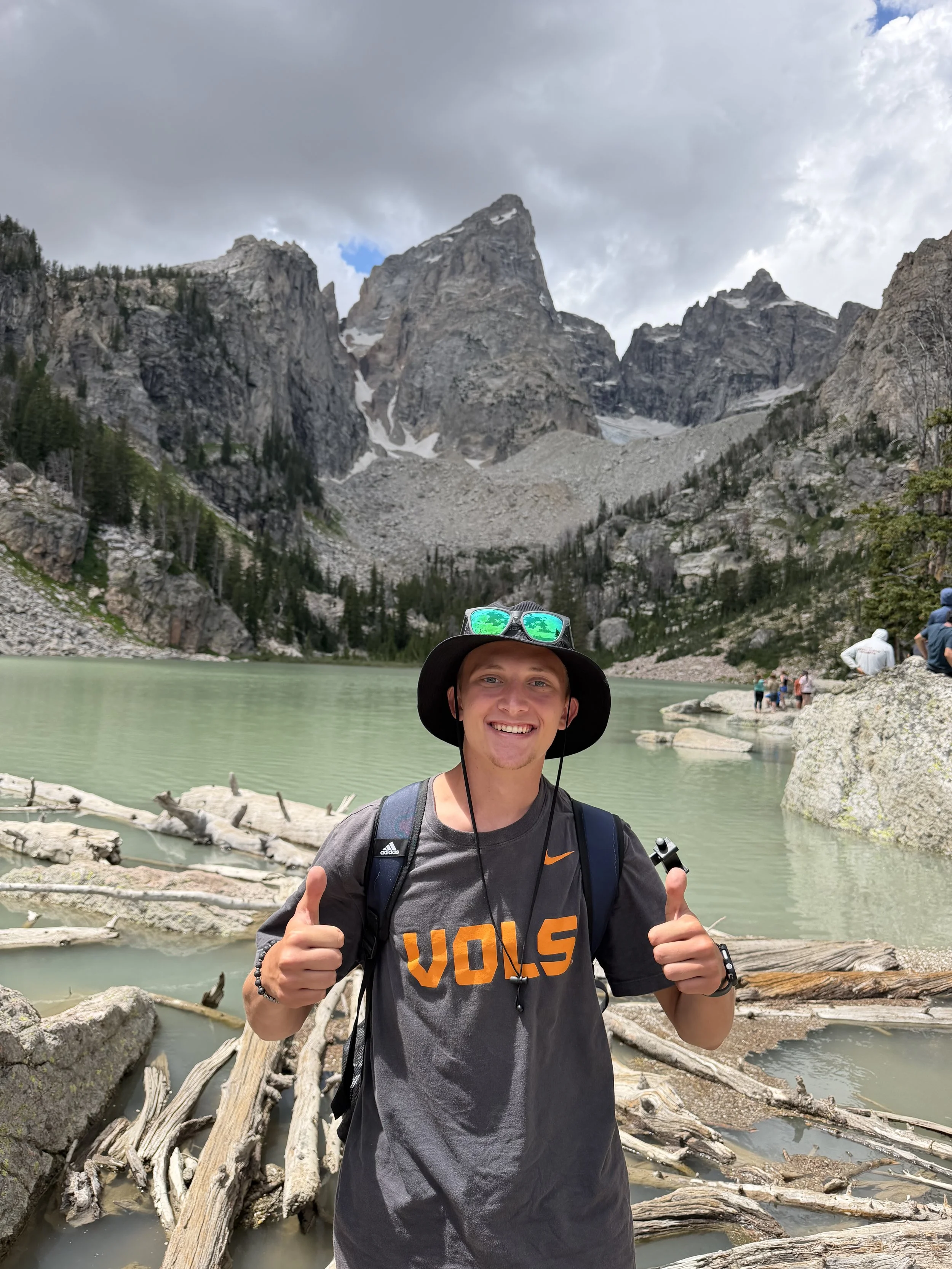 Smiling man hiking near a turquoise river with fallen logs, mountains, and cloudy sky in the background, giving a thumbs-up.