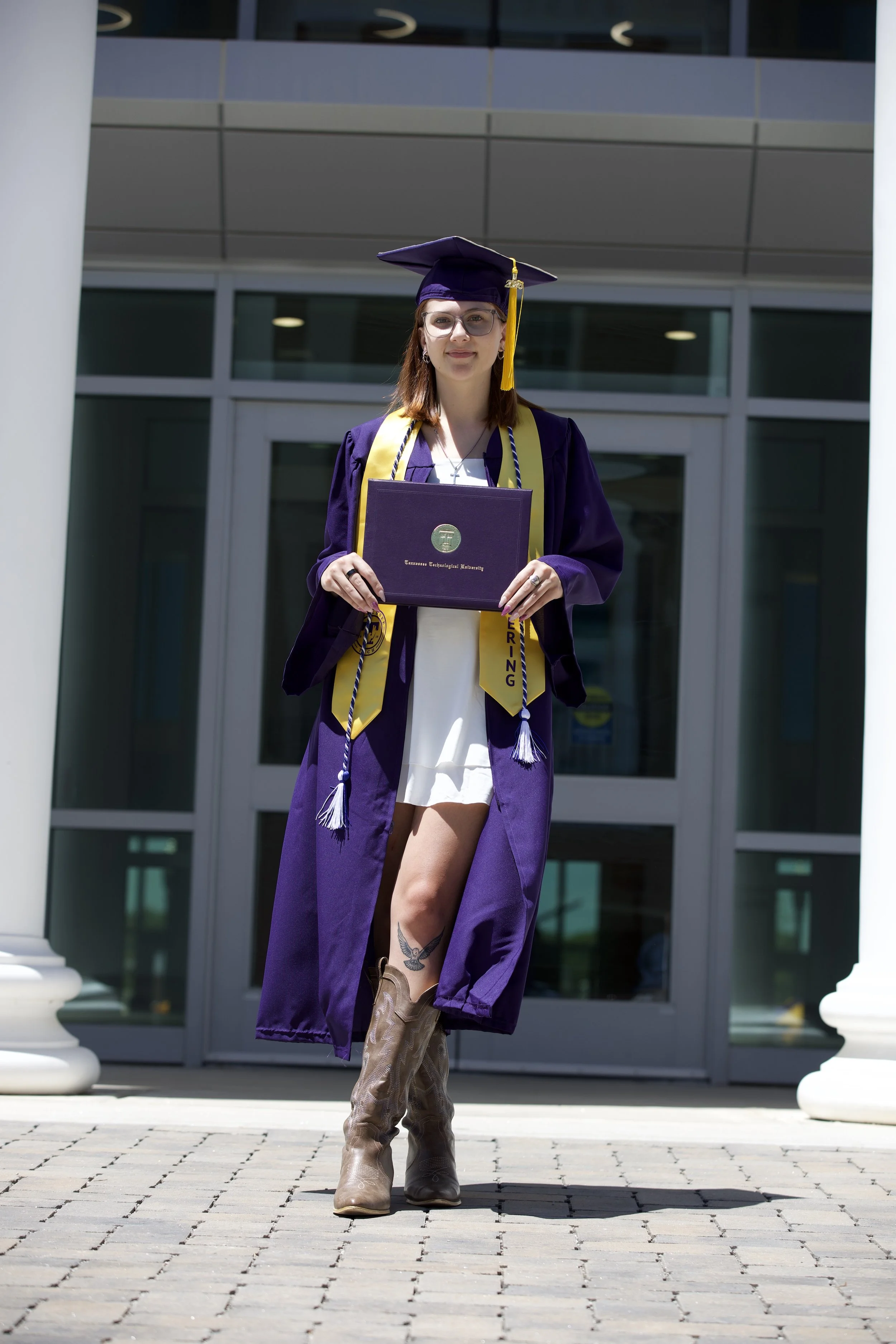 Young woman in a purple graduation gown and cap holding a diploma outside a building with large glass windows.