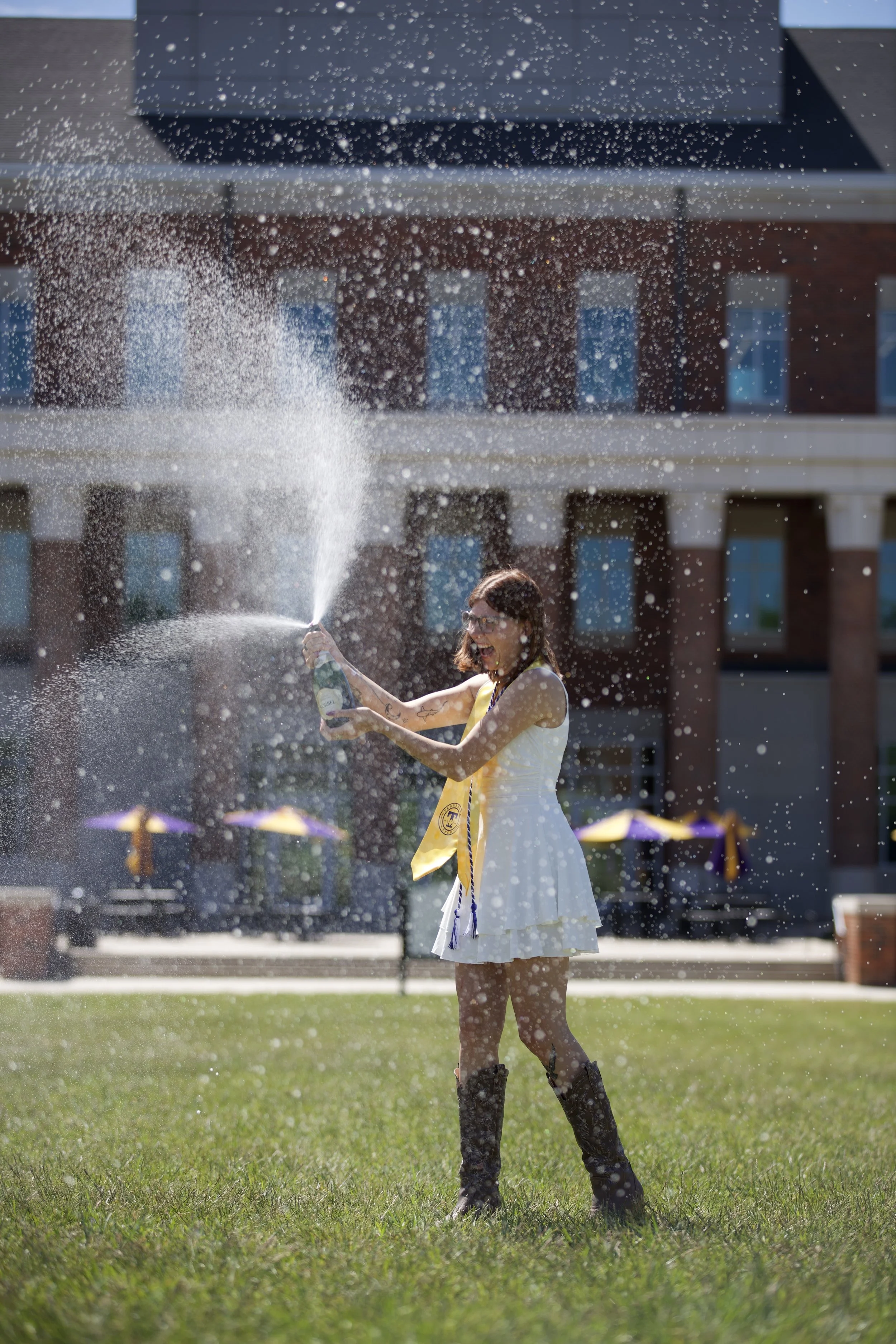 A woman with glasses, wearing a white dress and cowboy boots, is celebrating with a bottle of champagne during a graduation ceremony, standing on a grassy field with buildings in the background, as she sprays champagne into the air.