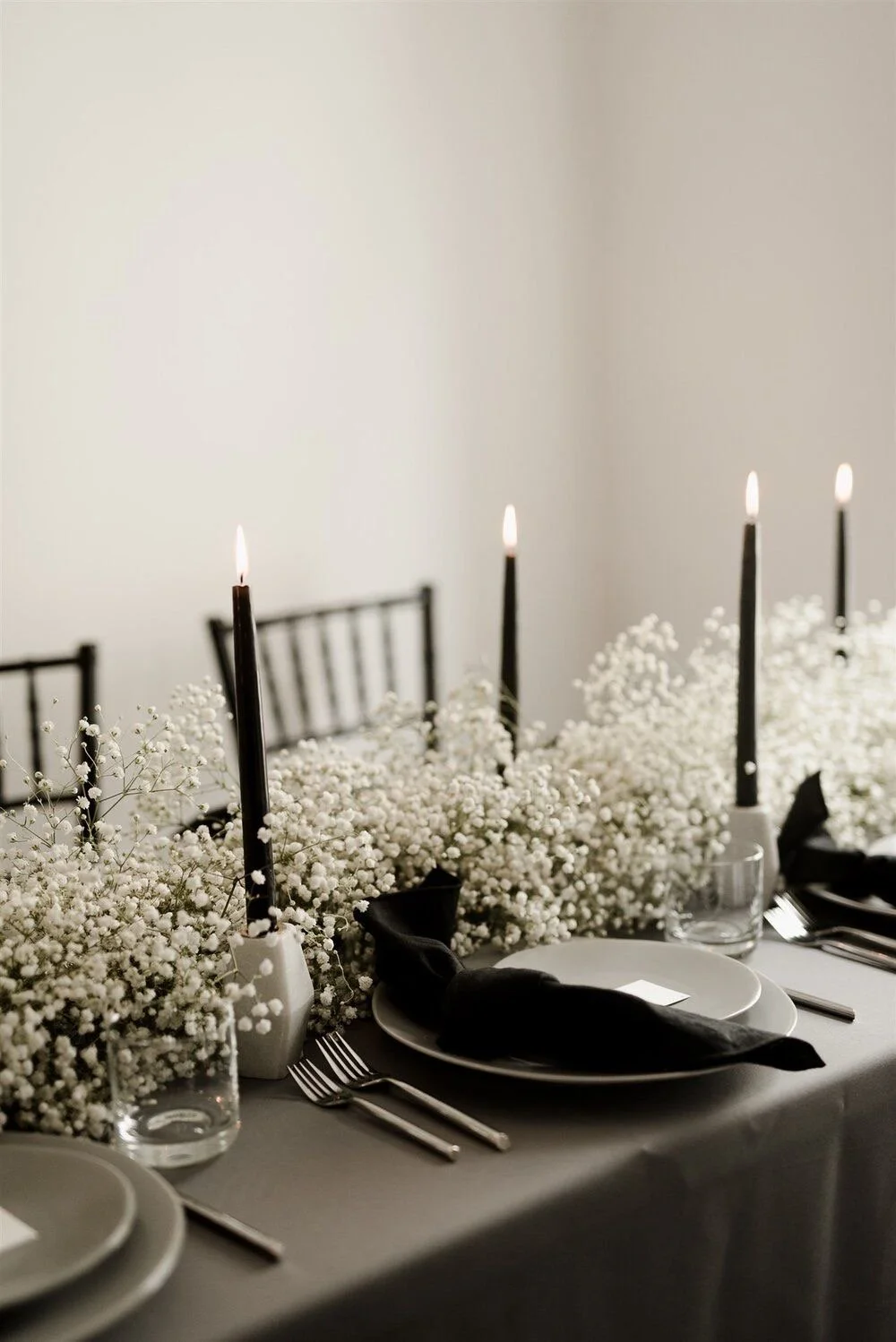 A dining table decorated with white baby's breath flowers, black candles, white plates, and silverware, set for a formal event.