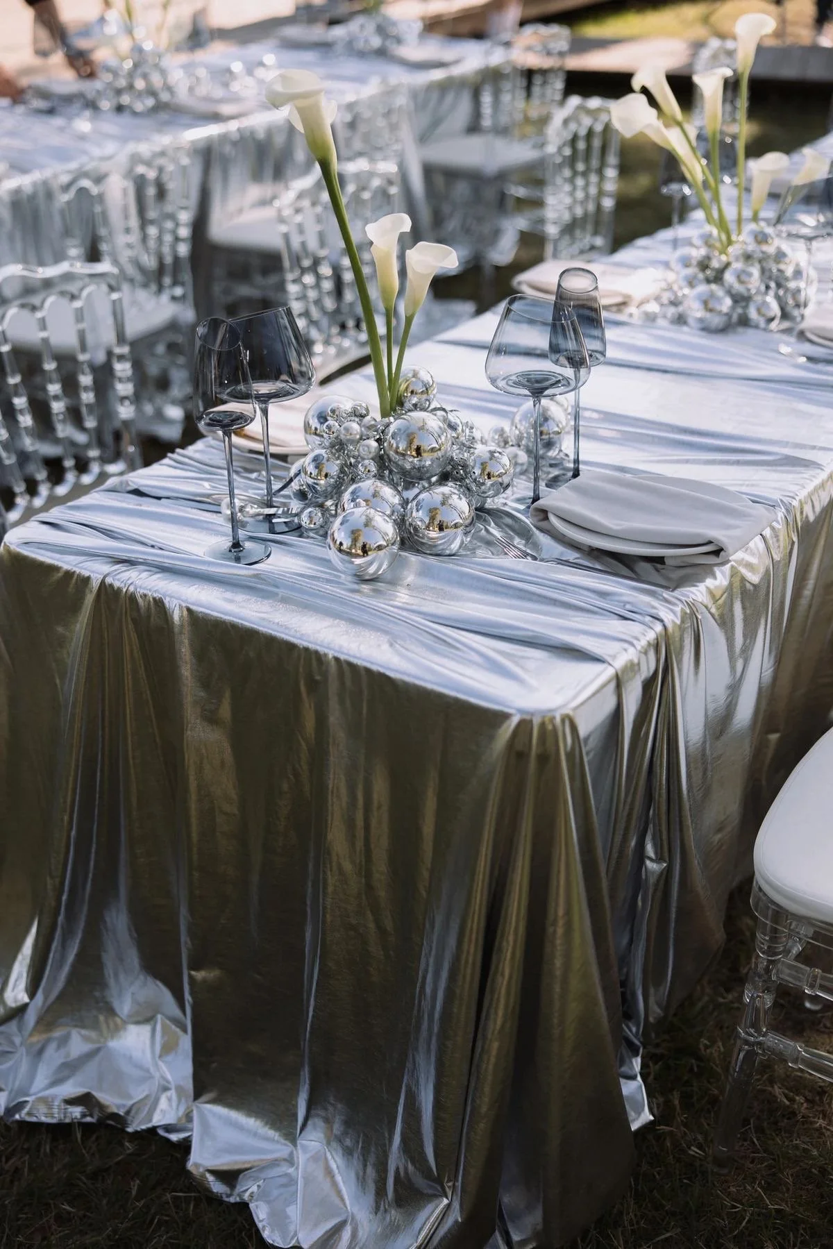 A dining table covered with a shiny silver tablecloth set for an event, decorated with a centerpiece of white calla lilies surrounded by shiny silver and gold balls, with white napkins and empty wine glasses all around.