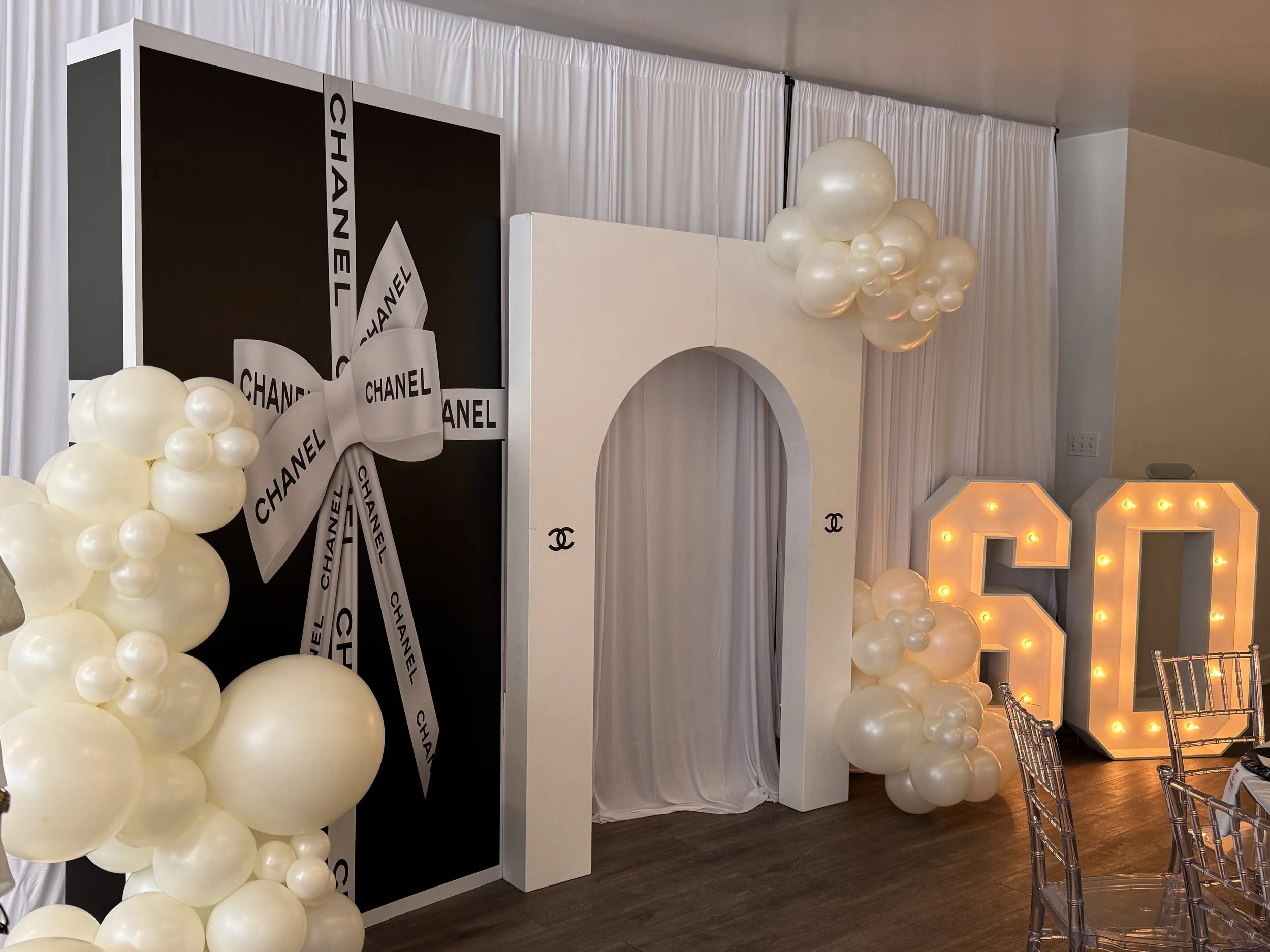 Decorative area with black and white Chanel-themed backdrop, white balloon clusters, a large illuminated letter 'G', and elegant chairs, likely for a celebration or event. Philadelphia event planner, Philadelphia birthday planner. Philadelphia decor.
