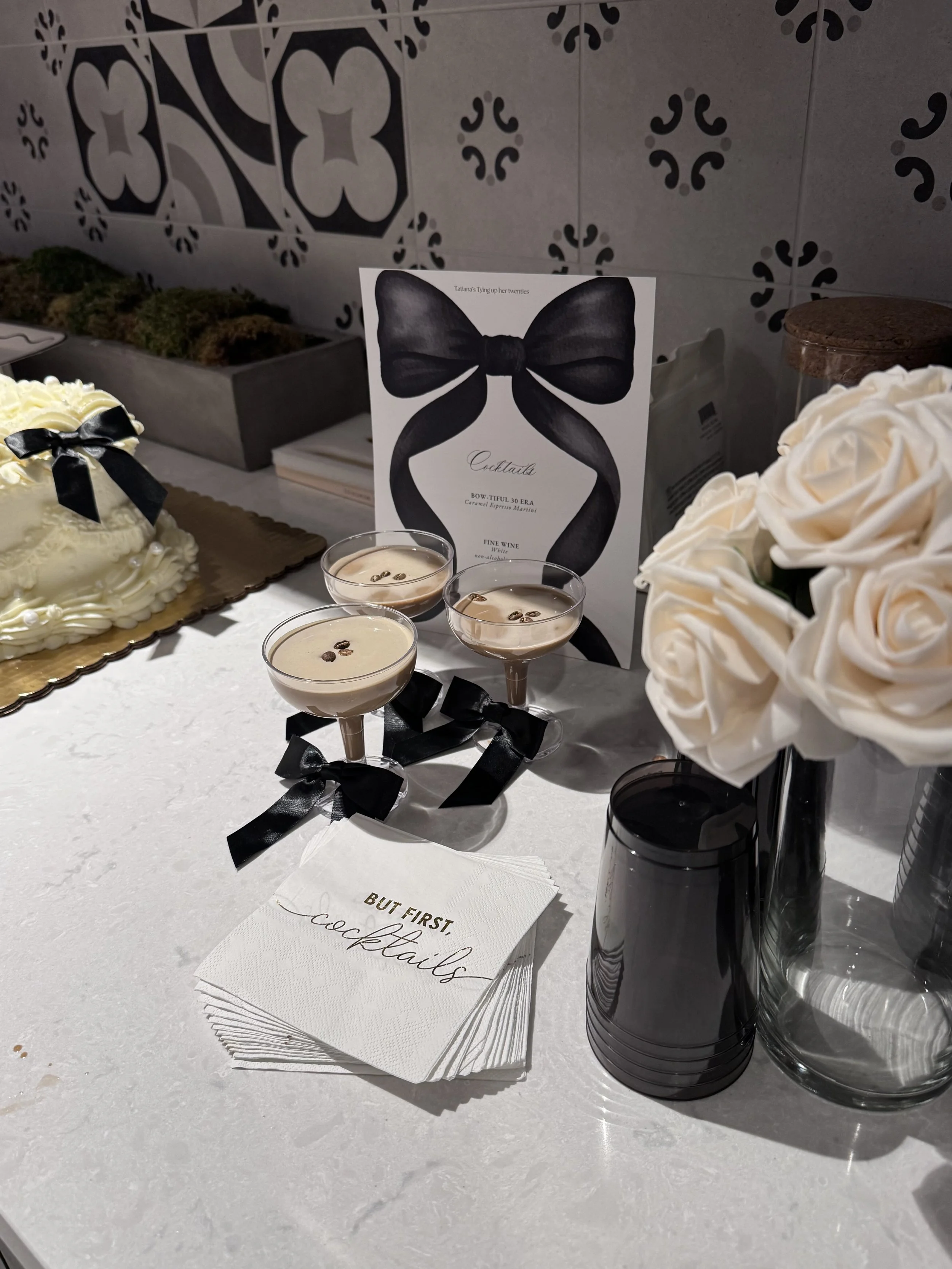 A table with white napkins, some text on a napkin that says 'BUT FIRST coffee,' three beige-colored cups of coffee with coffee beans on top, a bouquet of white roses in a vase, a card with a black bow illustration and the text 'Cocktails,' a small jar with a cork lid, and a gray rectangular box with plants inside. The background features a decorative tile wall.