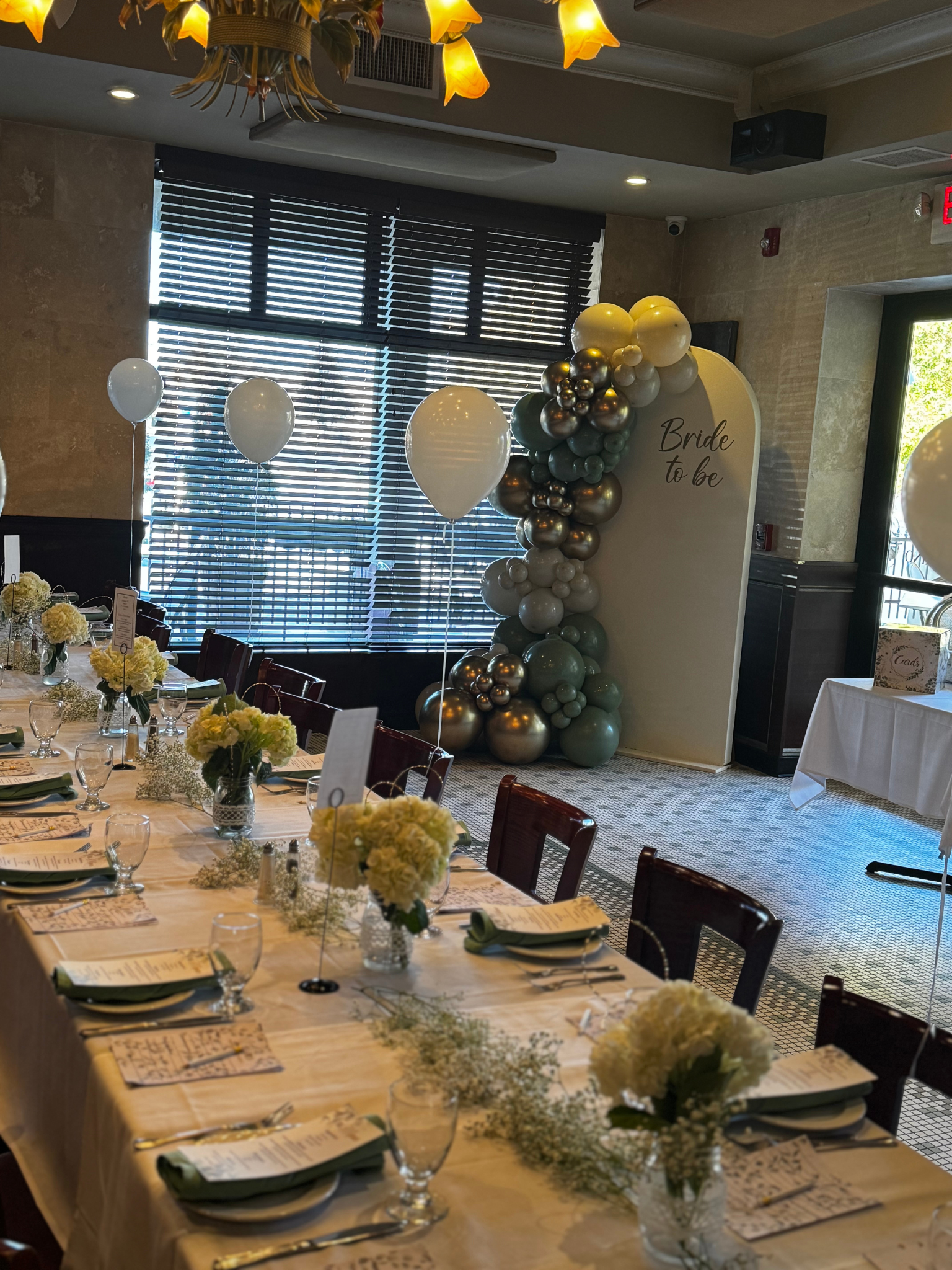 Decorated banquet table at a wedding reception with a flower arrangement, papers, glasses, and silverware, with a balloon arch and a 'Bride to be' sign in the background.Philadelphia event planner, Philadelphia birthday planner. Philadelphia decor