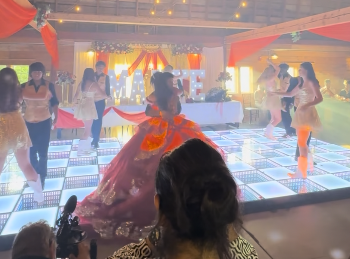 People dancing at a celebration with a woman in an elaborate gown on a lit dance floor, decorated with red drapery and floral arrangements, and large illuminated letters spelling 'MAME' in the background.