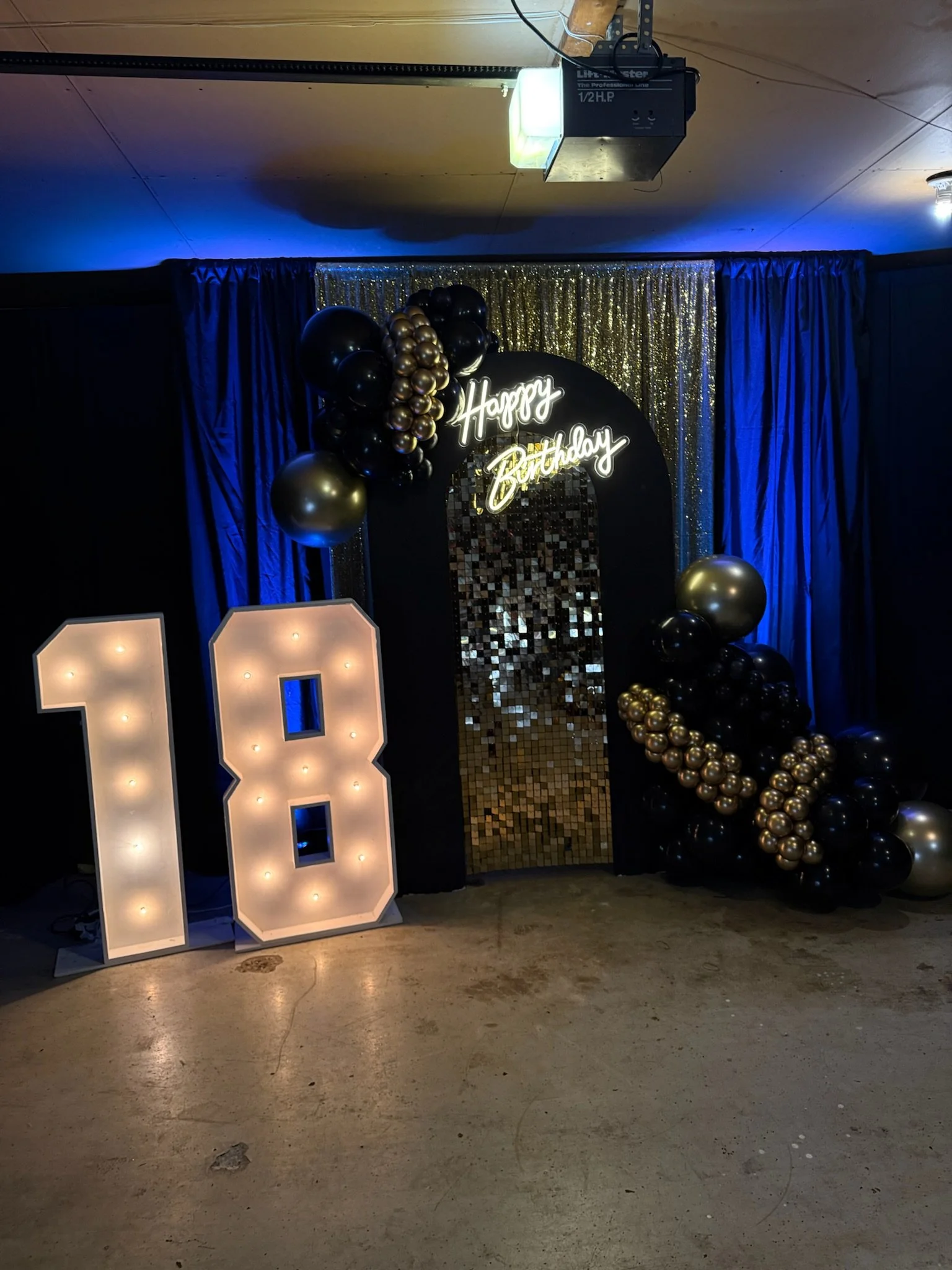 Decorative birthday party backdrop with illuminated number 8, gold and black balloons, a neon sign saying 'Happy Birthday,' golden sequin curtain, and a mirror mosaic panel.