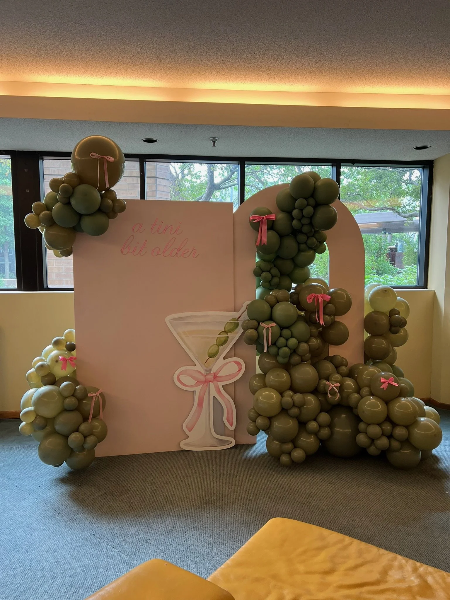 Decorative display with green balloons arranged around a white board with pink ribbon accents and the phrase 'a tini bit older' written on it.
