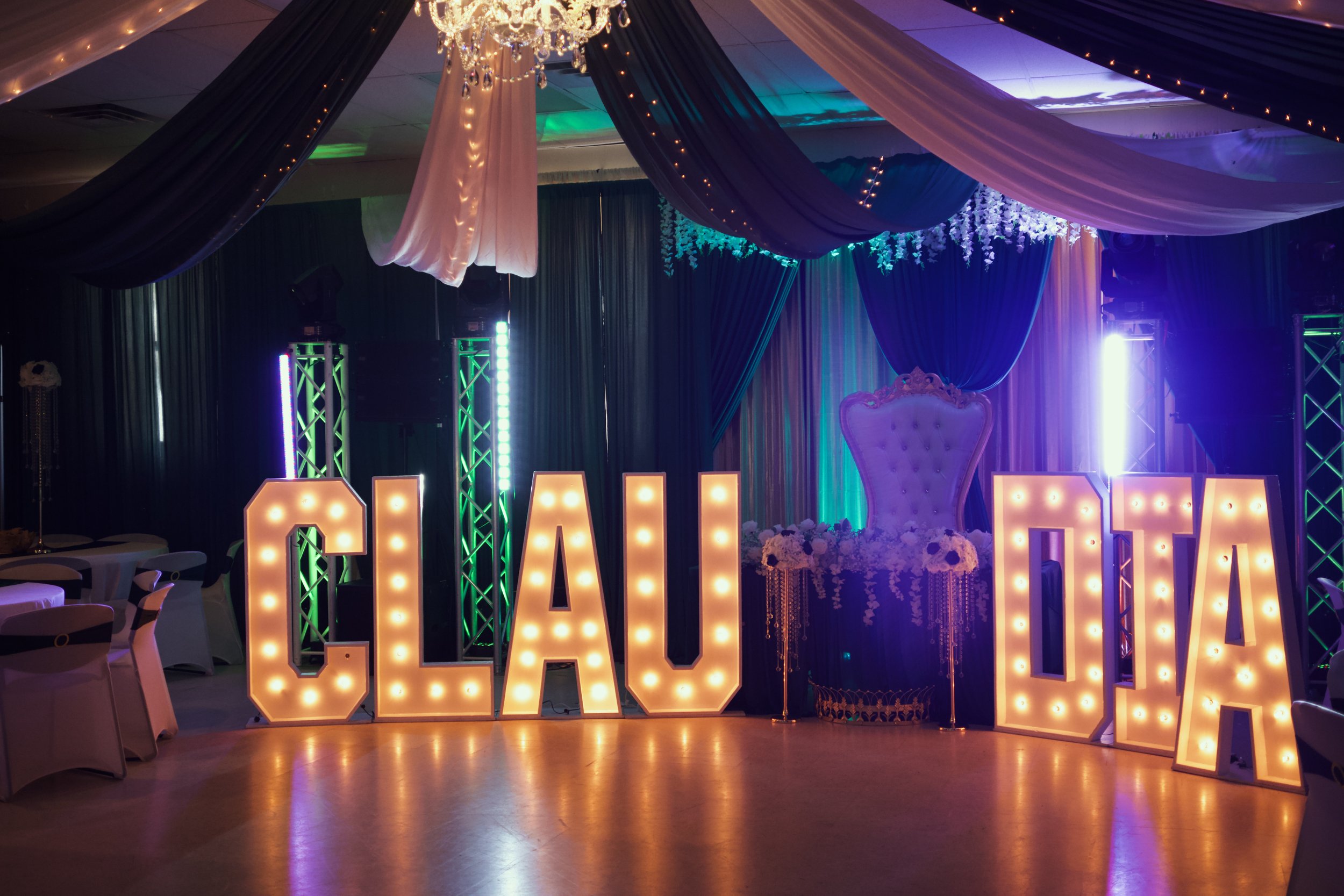 Decorated event stage with large illuminated letters, draped fabric, floral arrangements, and a chandelier, likely for a celebration or wedding reception.