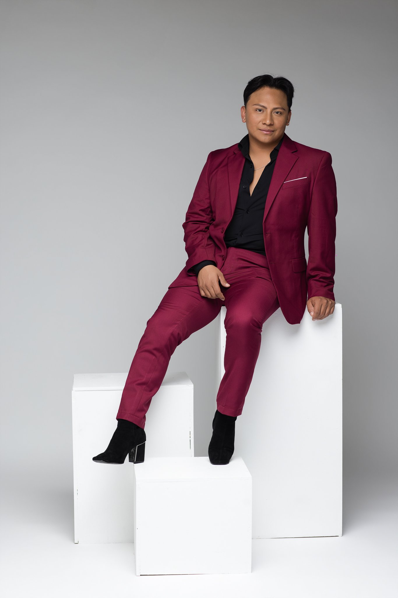 A person sitting on a white block, wearing a maroon suit, black shirt, and black boots, posing against a gray background.
