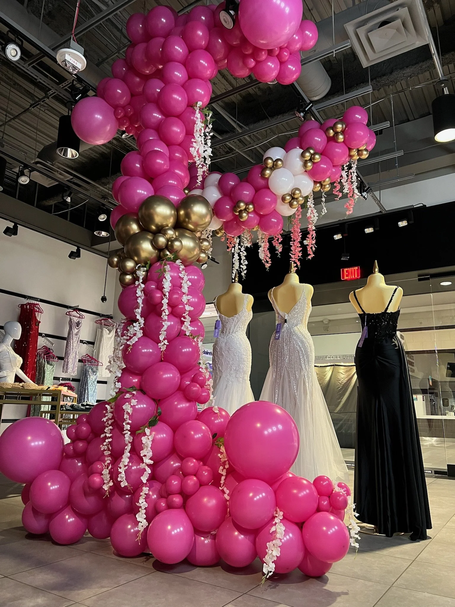 A display of pink, white, and gold balloons arranged in a large decorative cluster in a store that features mannequins wearing wedding dresses and dresses.