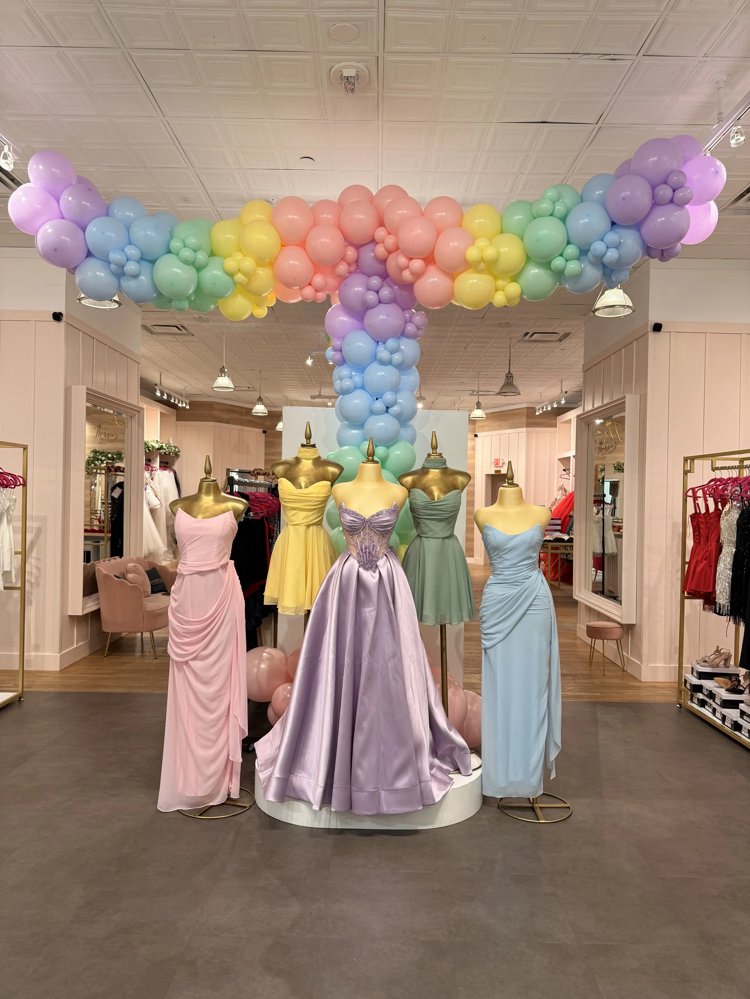 Display of mannequins dressed in pastel-colored dresses with a balloon sculpture overhead in a clothing store.