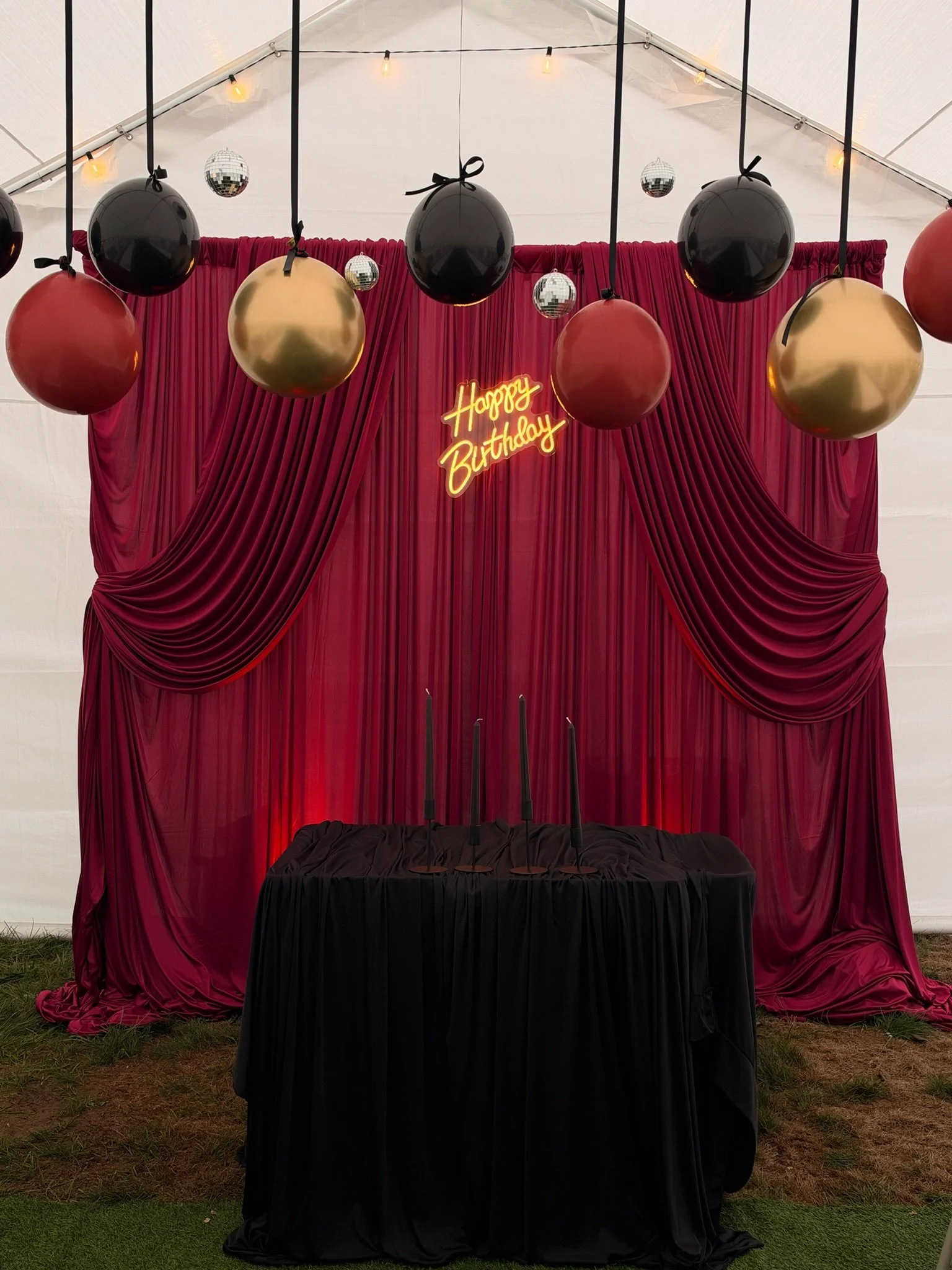 Decorative birthday party setup with a red curtain backdrop, black and gold balloons hanging from strings, and a neon sign that reads "Happy Birthday". There is a table covered with black cloth, and two black candles on it. Small disco balls and stri
