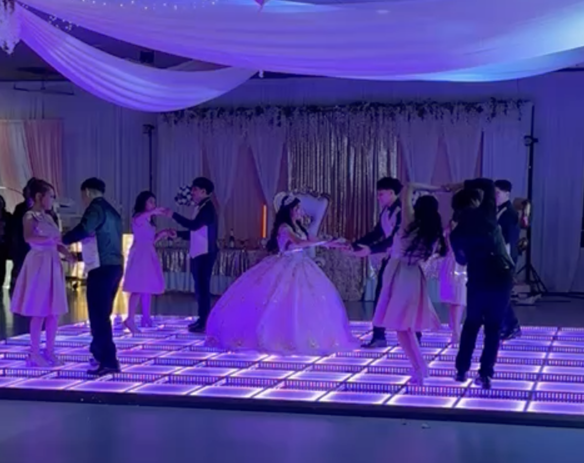 A group of children dancing on an illuminated dance floor at a birthday party or celebration, with a girl in a pink dress and crown in the center, surrounded by other kids.