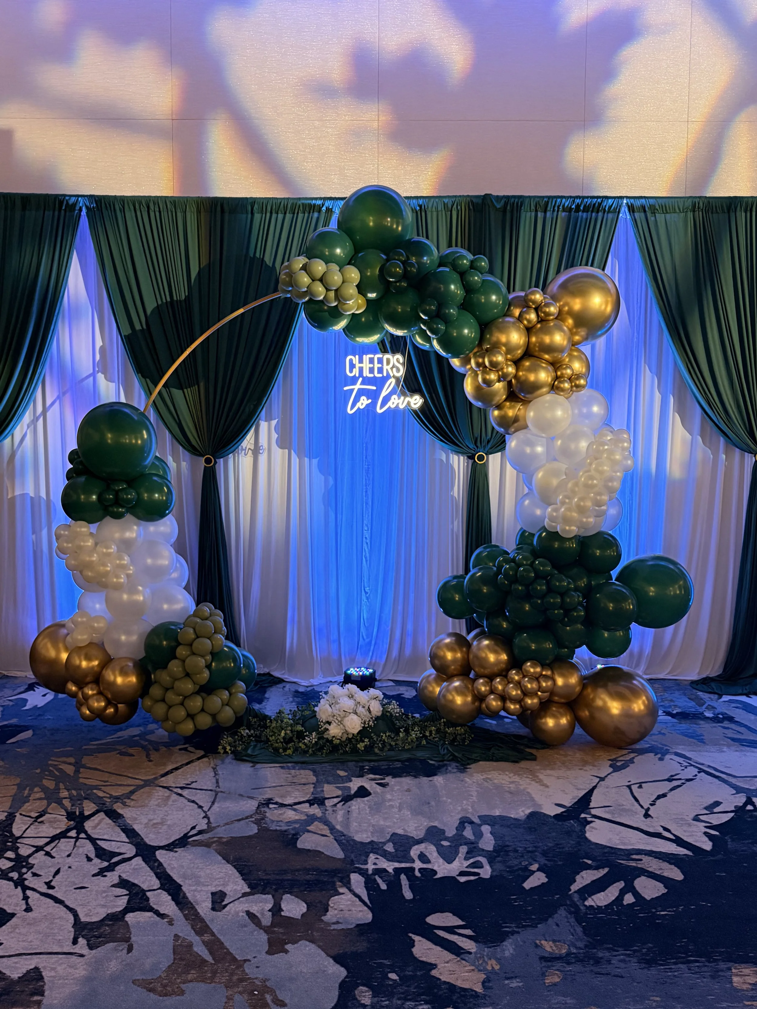 Decorative backdrop with dark green drapes, white sheer curtains, and gold, green, and white balloons arranged in an arch, centered around a neon sign that says 'Cheers to Love'; floral arrangement at the base.
