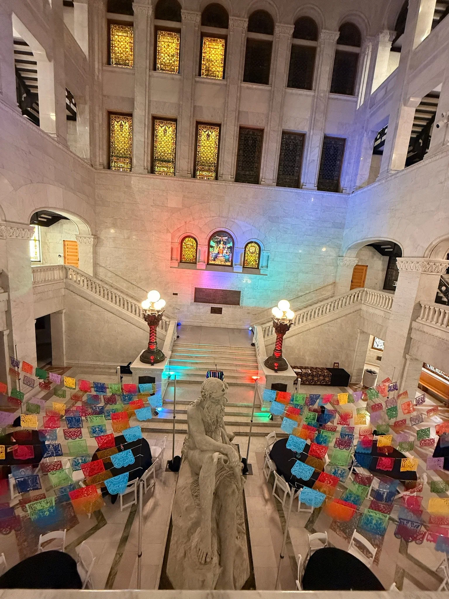 Interior of a grand, marble staircase hall with colorful papel picado decorations, stained glass windows, and a marble statue in the center.