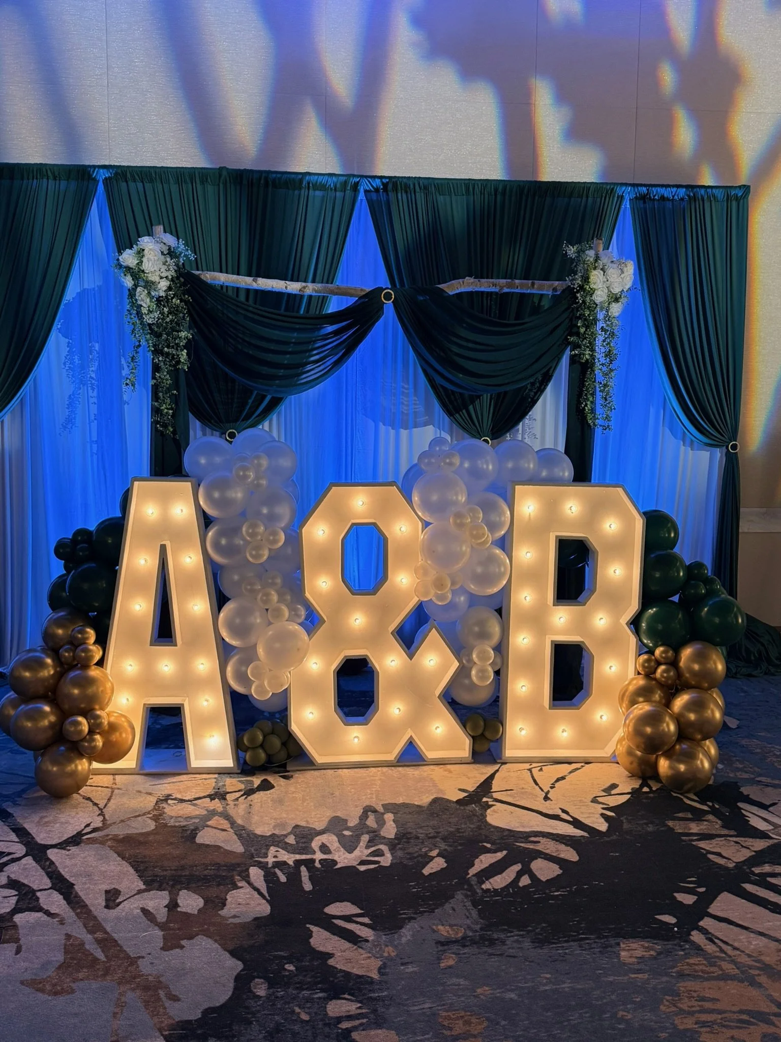 Decorative backdrop with dark green and white curtains, floral arrangements, balloons, and illuminated 'LOVE' letters at a celebration or event.