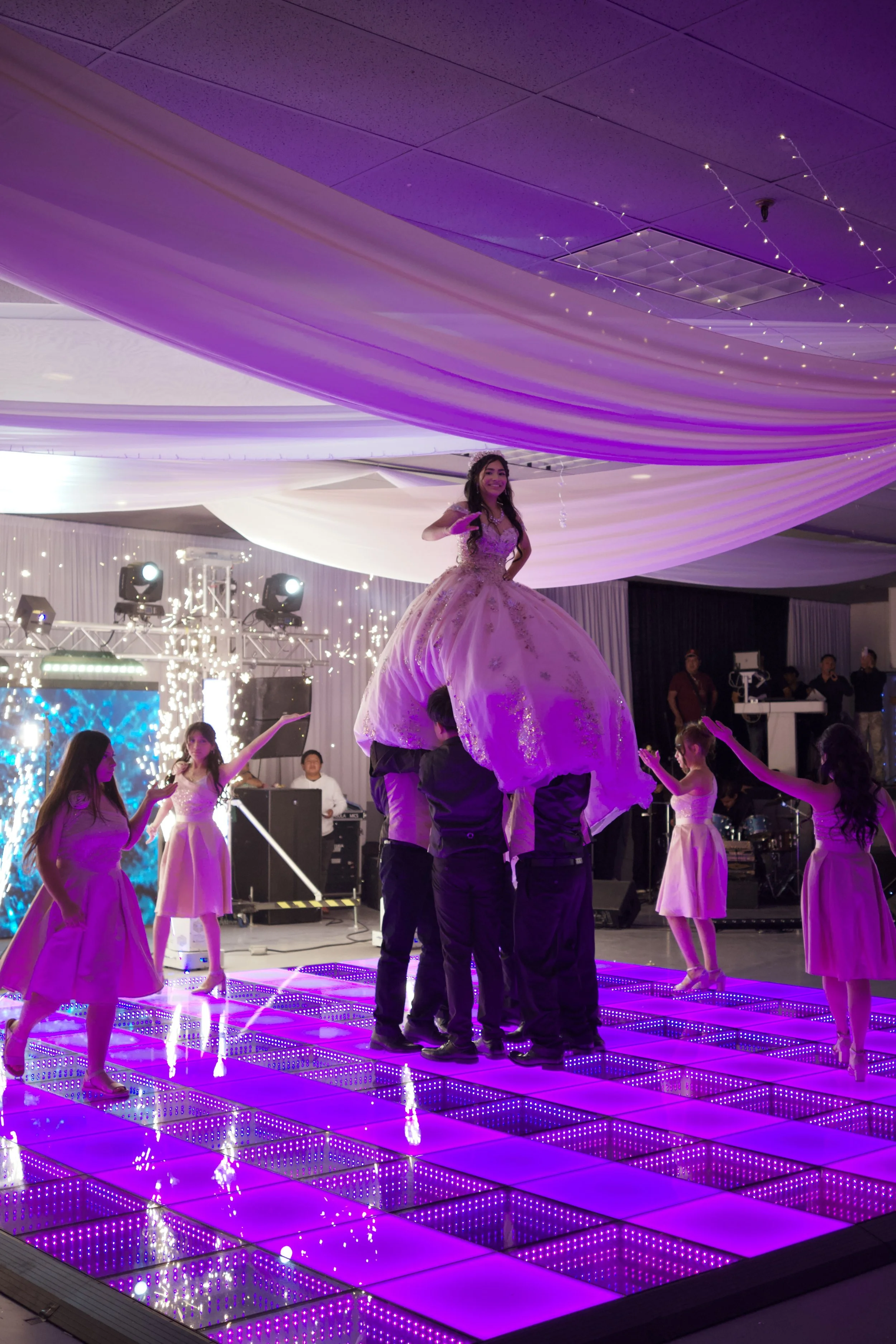 A girl in a fancy dress stands on a raised platform with purple LED lights, surrounded by young dancers in pink dresses, with a band and event lights in the background, during an indoor celebration or party.