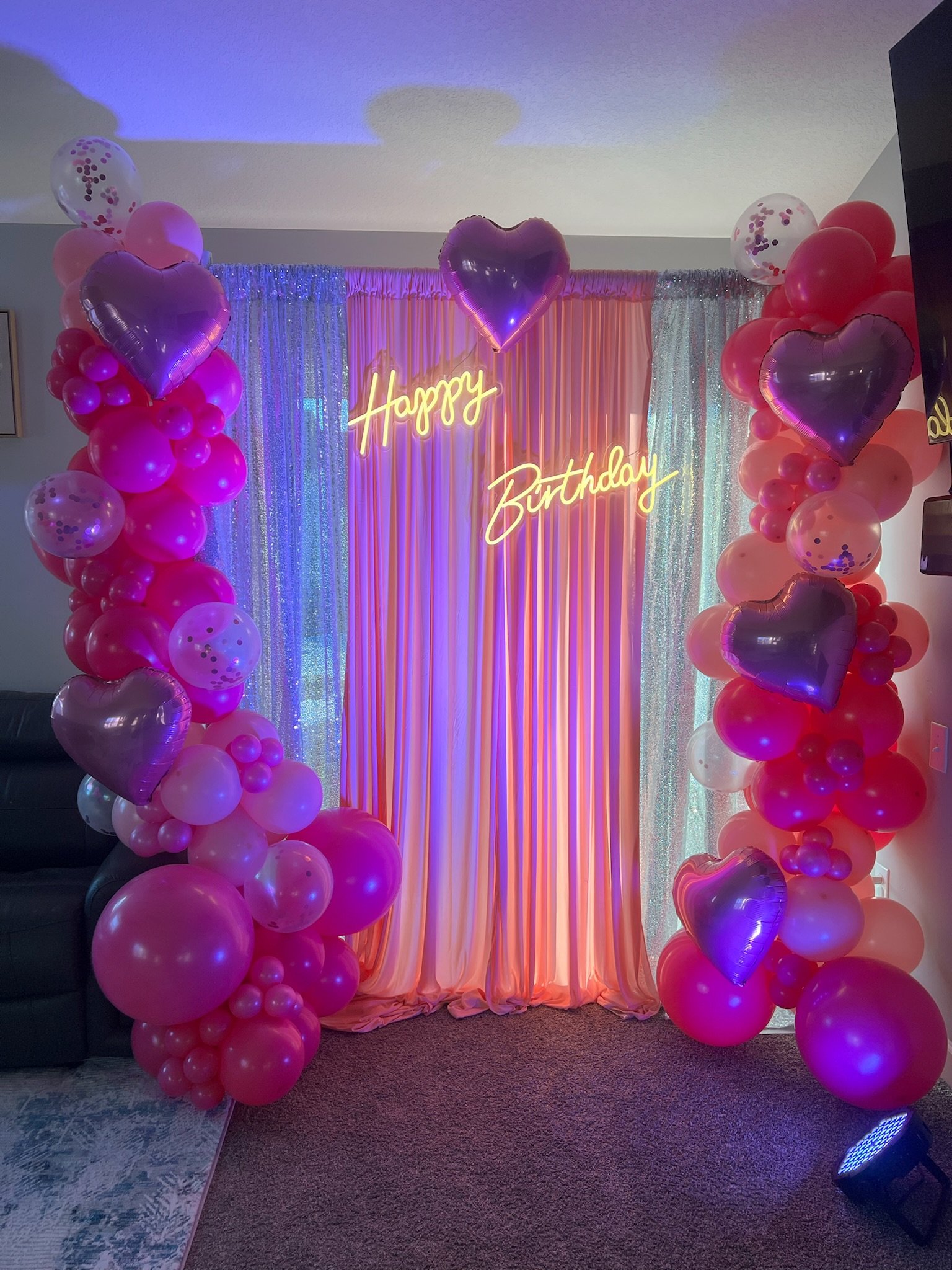Colorful birthday decoration with balloons, pink curtain backdrop, and neon sign that says 'Happy Birthday'.