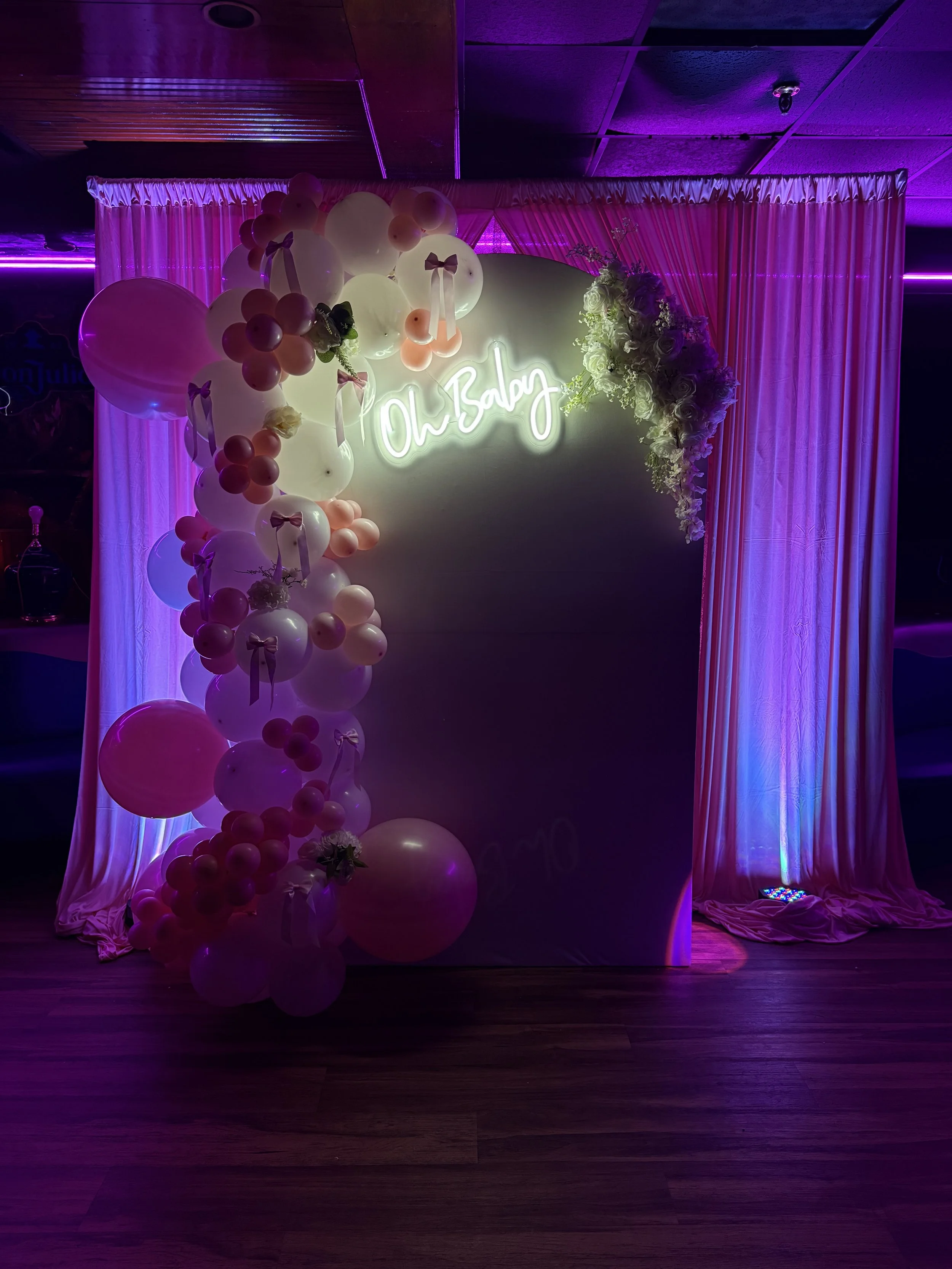 Decorative backdrop with pink and white balloons, pink curtains, white flowers, and neon sign that reads 'Oh Baby' in a dimly lit setting with purple and pink lighting.
