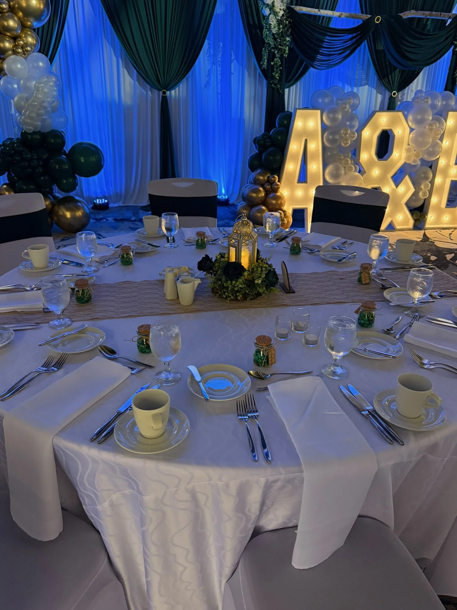 Table set for a celebration with white tablecloth, glassware, cups, plates, and utensils. Decorated with small jars and a lantern with candles in the center. In the background, large illuminated letters spell out some words, surrounded by black, whit
