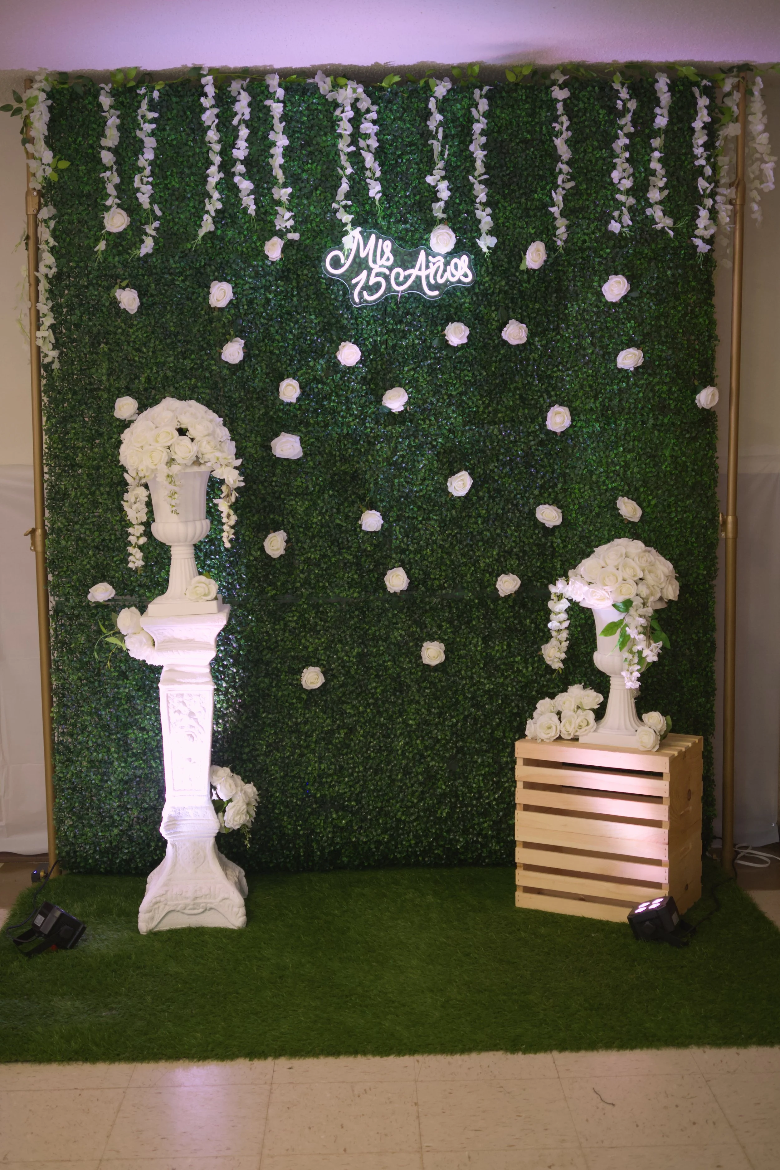 Green artificial hedge wall decorated with white hanging flowers and roses, illuminated sign reading 'Mrs 150 Anos', white flower arrangements on tall white stands and a wooden crate, black spotlights on a grassy floor, ceiling with pinkish hue.