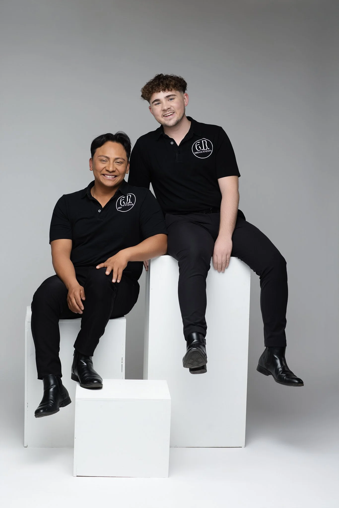 Two young men dressed in black, sitting and standing on white blocks against a plain gray background, smiling for the camera. Both wear black polo shirts with a logo that reads 'G.D. Dream Events,' and black boots.