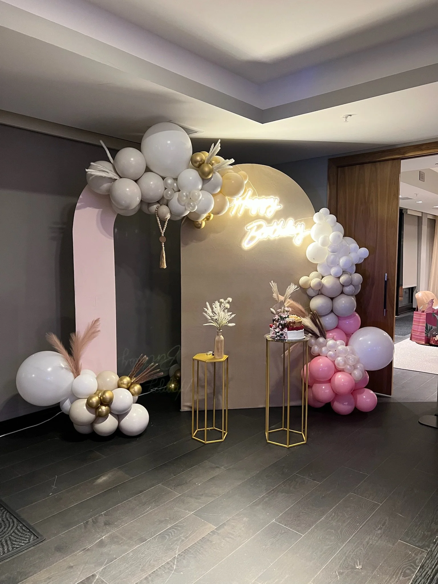 Decorative balloon arch with white, pink, and gold balloons surrounding a backdrop with neon sign that reads 'Tiffany Baby' and pink and white floral arrangements at the base.