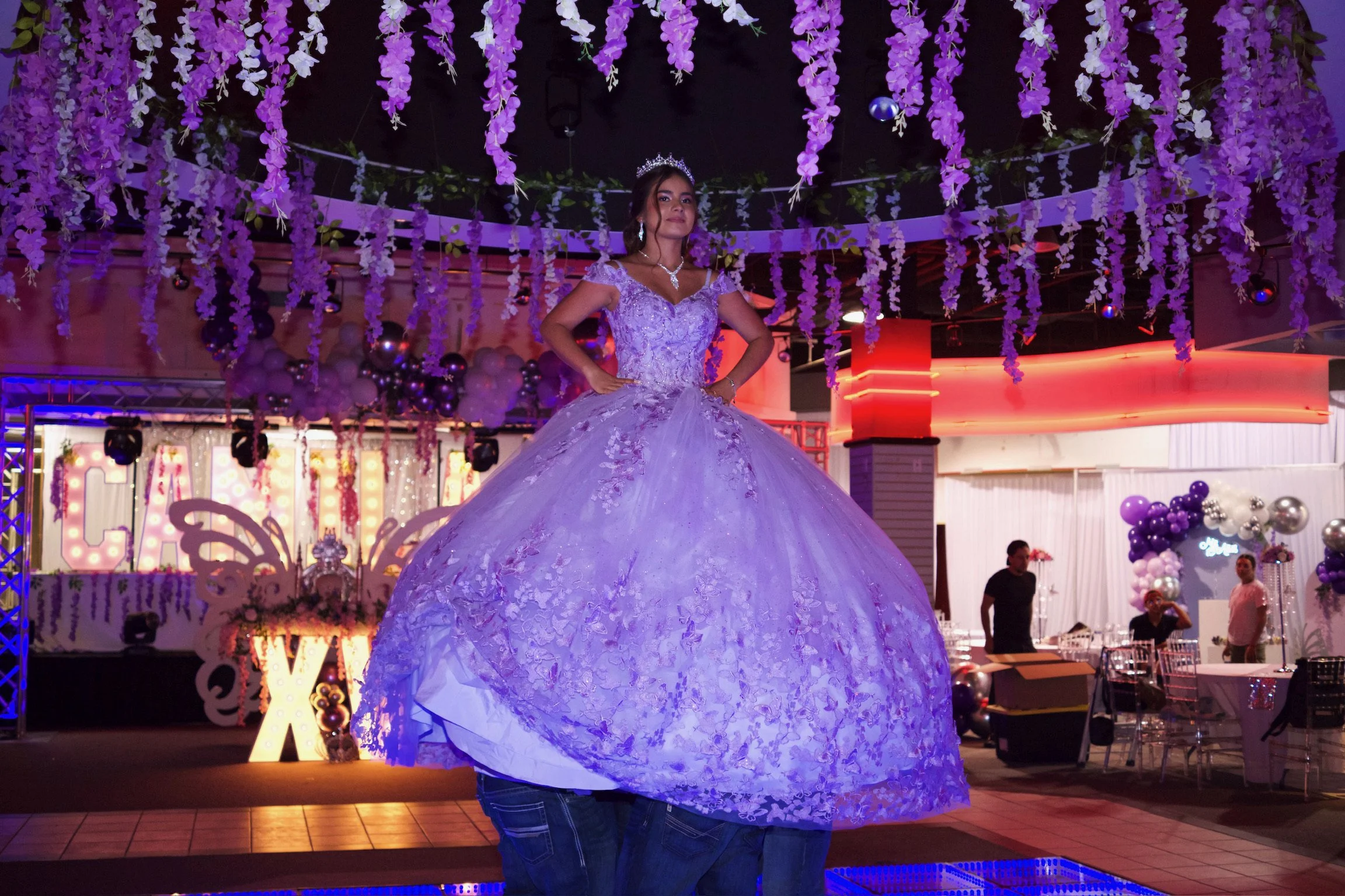 A woman posing in a lavender ball gown with lace details at a celebration or event, decorated with purple and white floral arrangements and glowing colored lights, in a venue with a festive atmosphere.