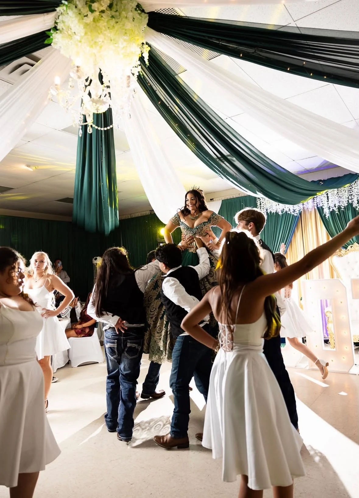 A lively celebration with people dancing and a woman being lifted on the shoulders of others in a decorated venue with green and white drapes and ornate ceiling decorations.