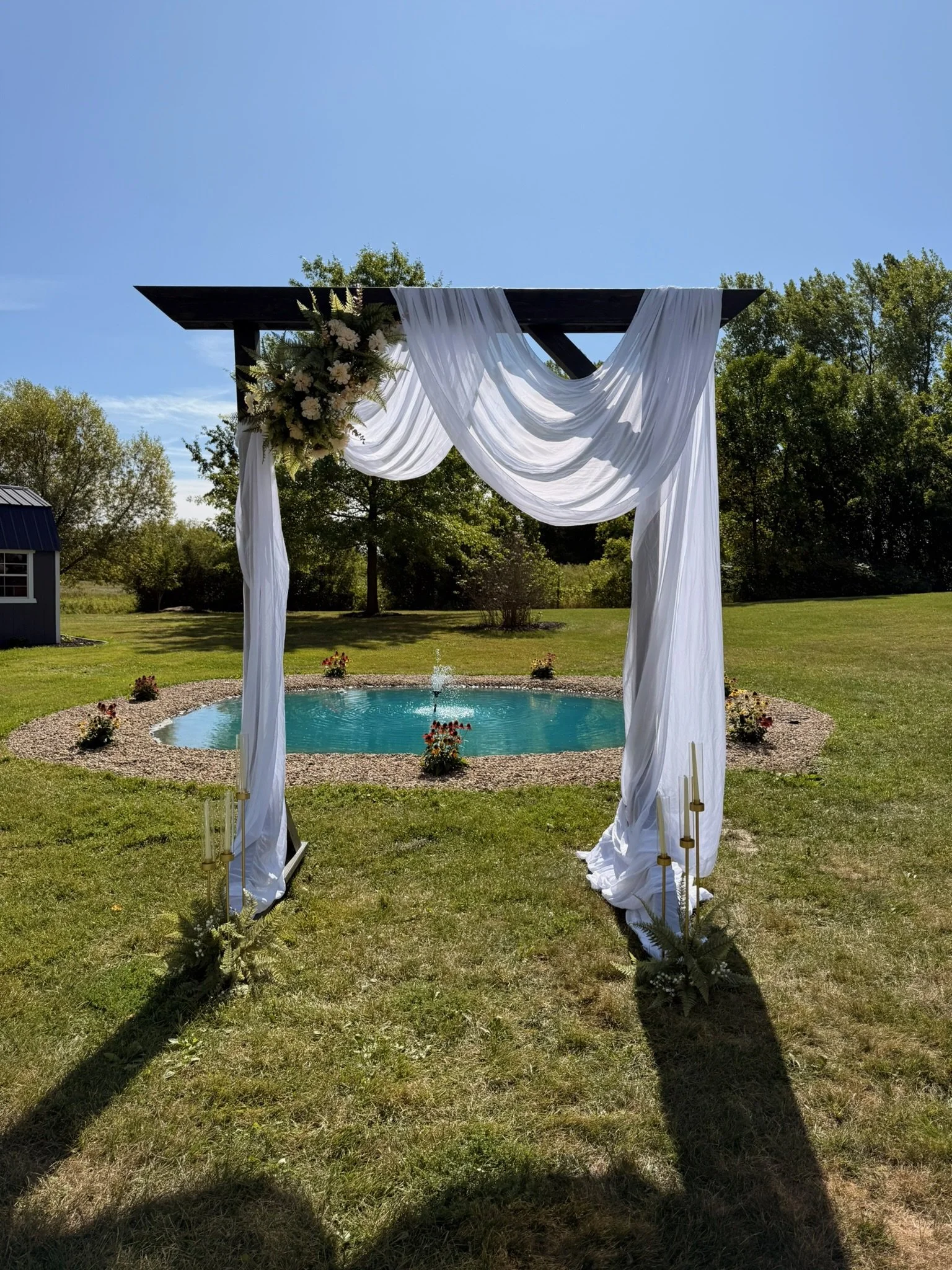 Outdoor wedding arch decorated with white drapery and flowers, situated over a small pond on a grassy lawn with trees in the background and a clear blue sky.