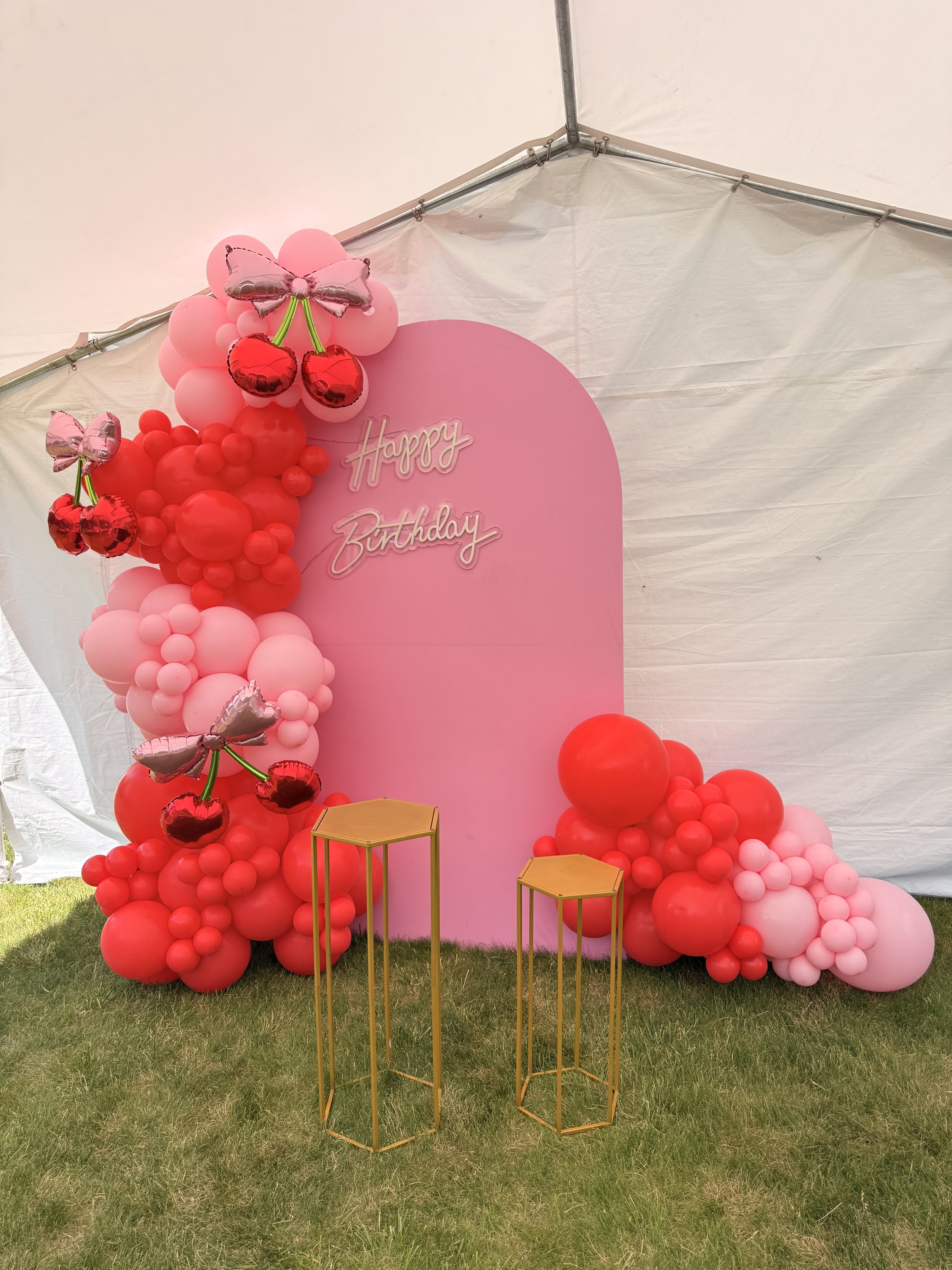 Decorative pink backdrop with balloons and a "Happy Birthday" sign, set up for a celebration. There are pink, red, and light pink balloons arranged in clusters along with silver cherry-shaped balloons with green stems and silver butterfly-shaped ball