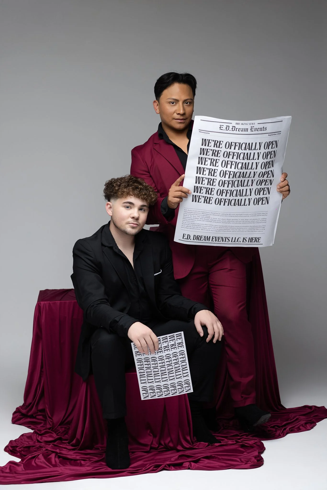 Two young men, one seated and one standing, holding signs that say 'We're officially open,' with a gray background and burgundy fabric on the floor and furniture.