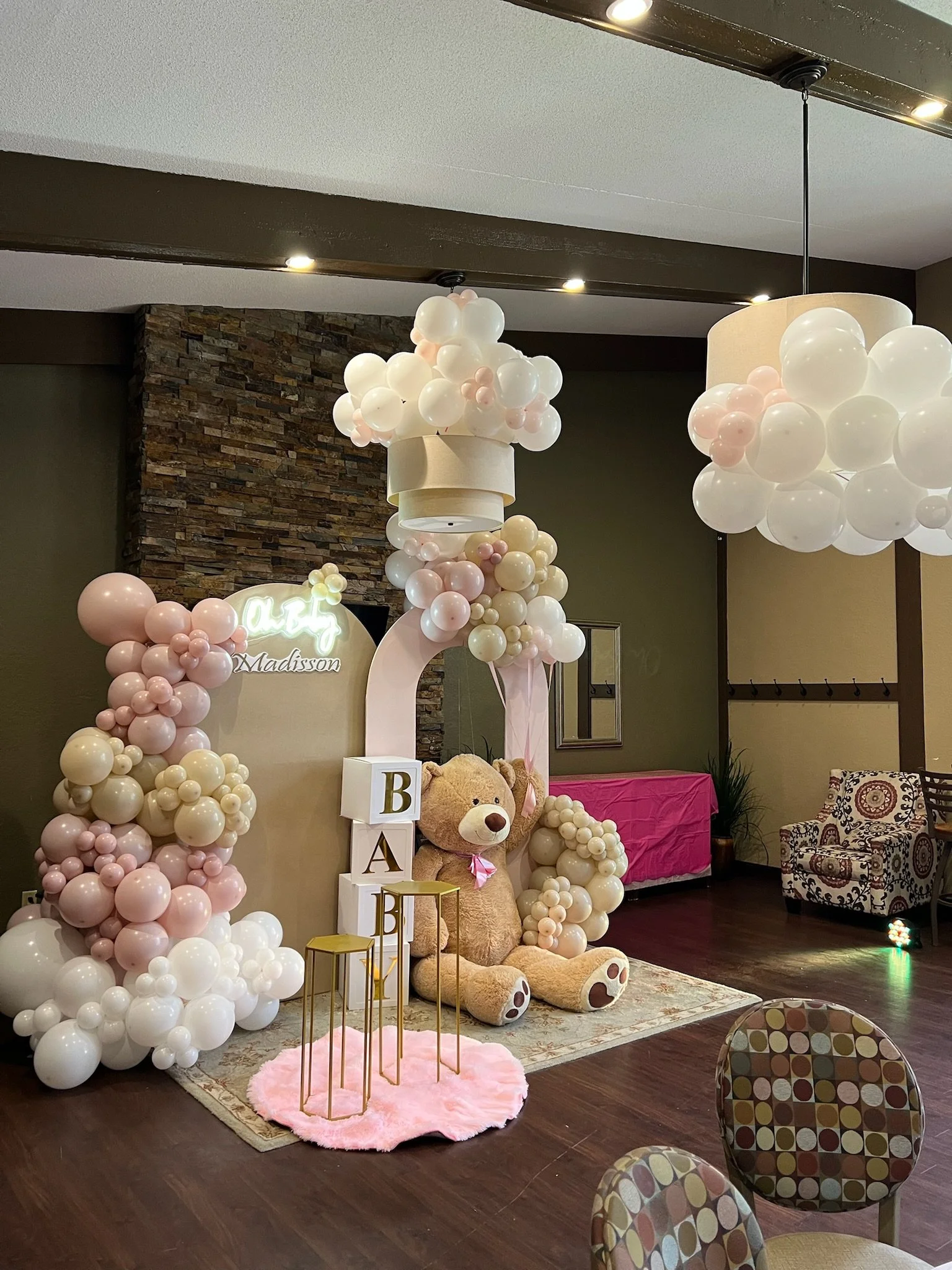 Decorative baby shower setup with white and pink balloons, a large teddy bear, a neon sign, and pink accents. There are chairs with a multicolored pattern and a pink fluffy rug on dark wood flooring.