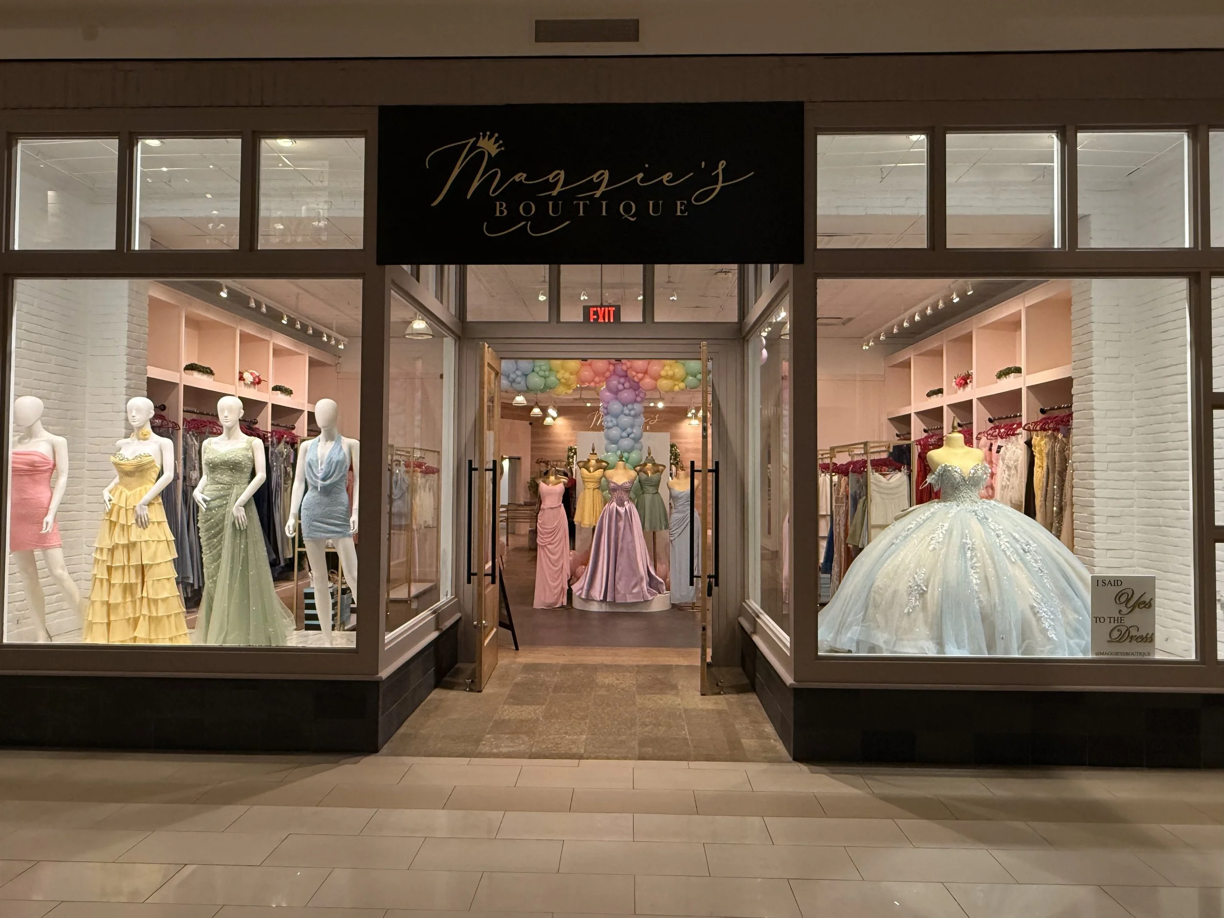 Storefront window of Maggie's Boutique displaying mannequins dressed in pastel-colored dresses, with shelves of accessories and decorations inside, and a balloon arch visible further inside.