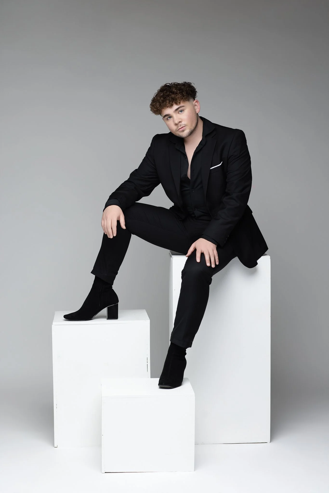 A young man with curly hair, dressed in a black suit, sitting on white blocks against a gray background.