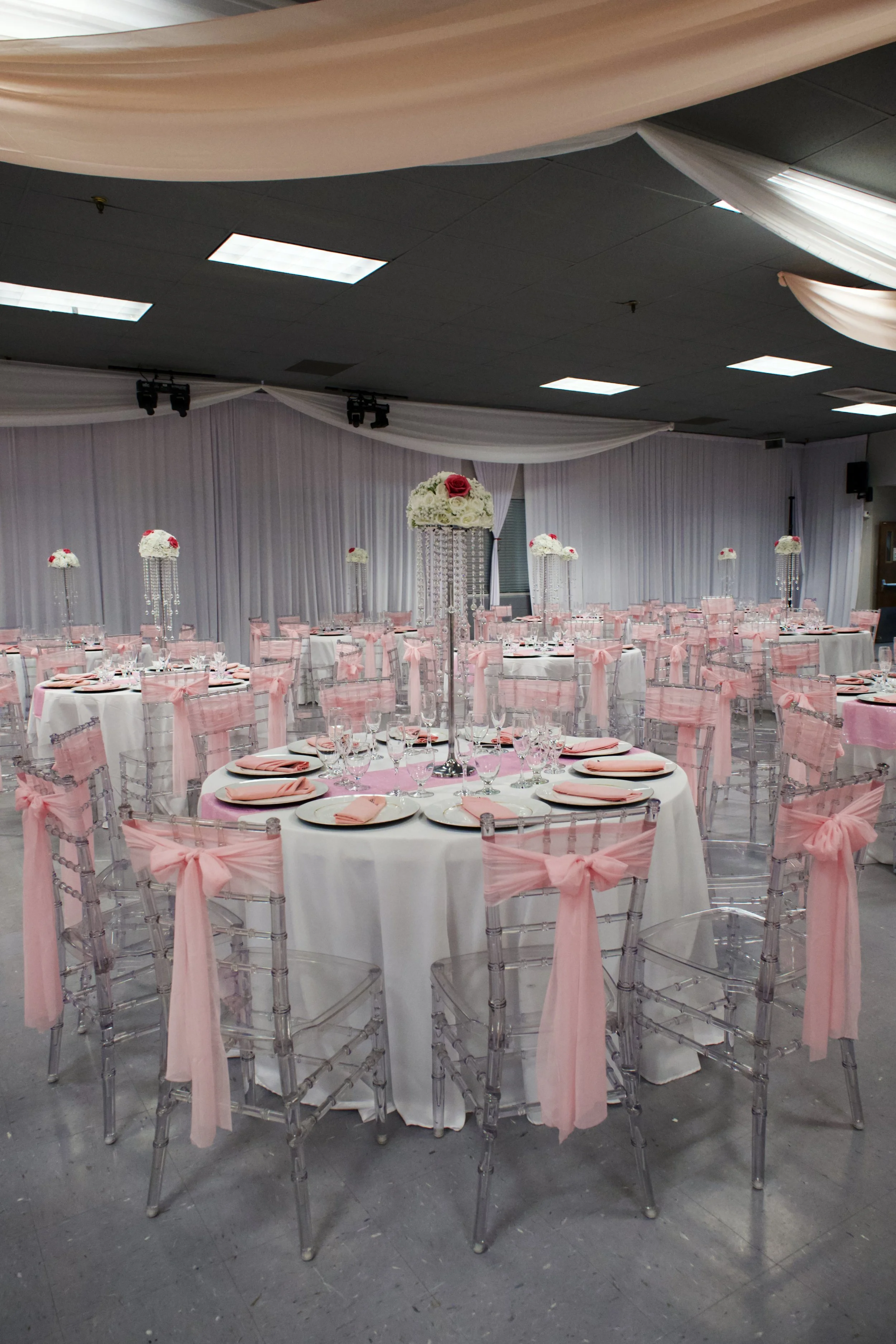 Wedding reception hall with round tables decorated with pink ribbons on transparent chairs, white tablecloths, pink napkins, and floral centerpieces with white and pink flowers.