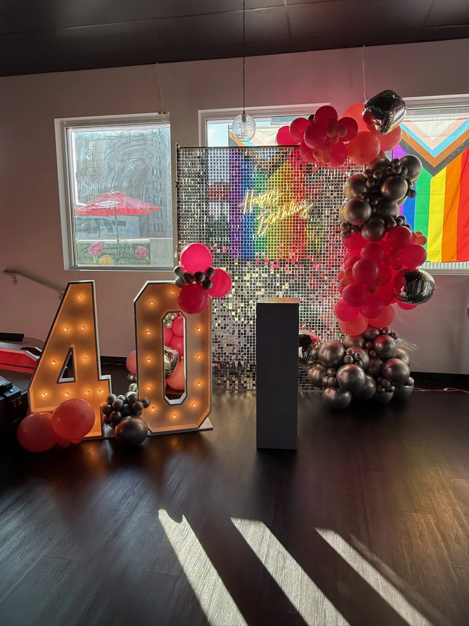 Decorative celebration setup with a large illuminated '40' sign, balloons in shades of pink, black, and silver, a metallic 'Happy Birthday' sign, and rainbow tape on the window in the background.