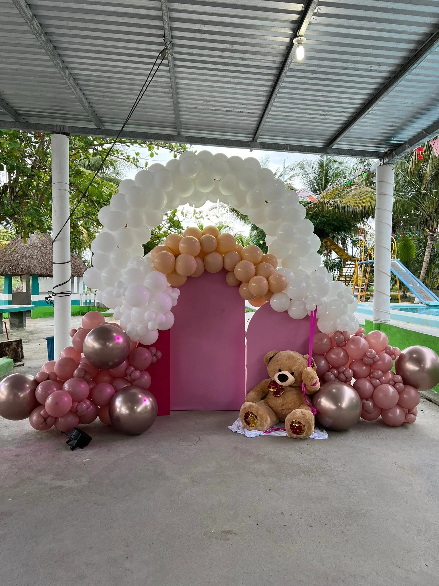 Decorative setup with pink and white balloons arranged in an arch and clusters, featuring a teddy bear with a bow tie sitting on a cloth in front, under a metal roof structure at an outdoor venue with trees and playground equipment in the background.