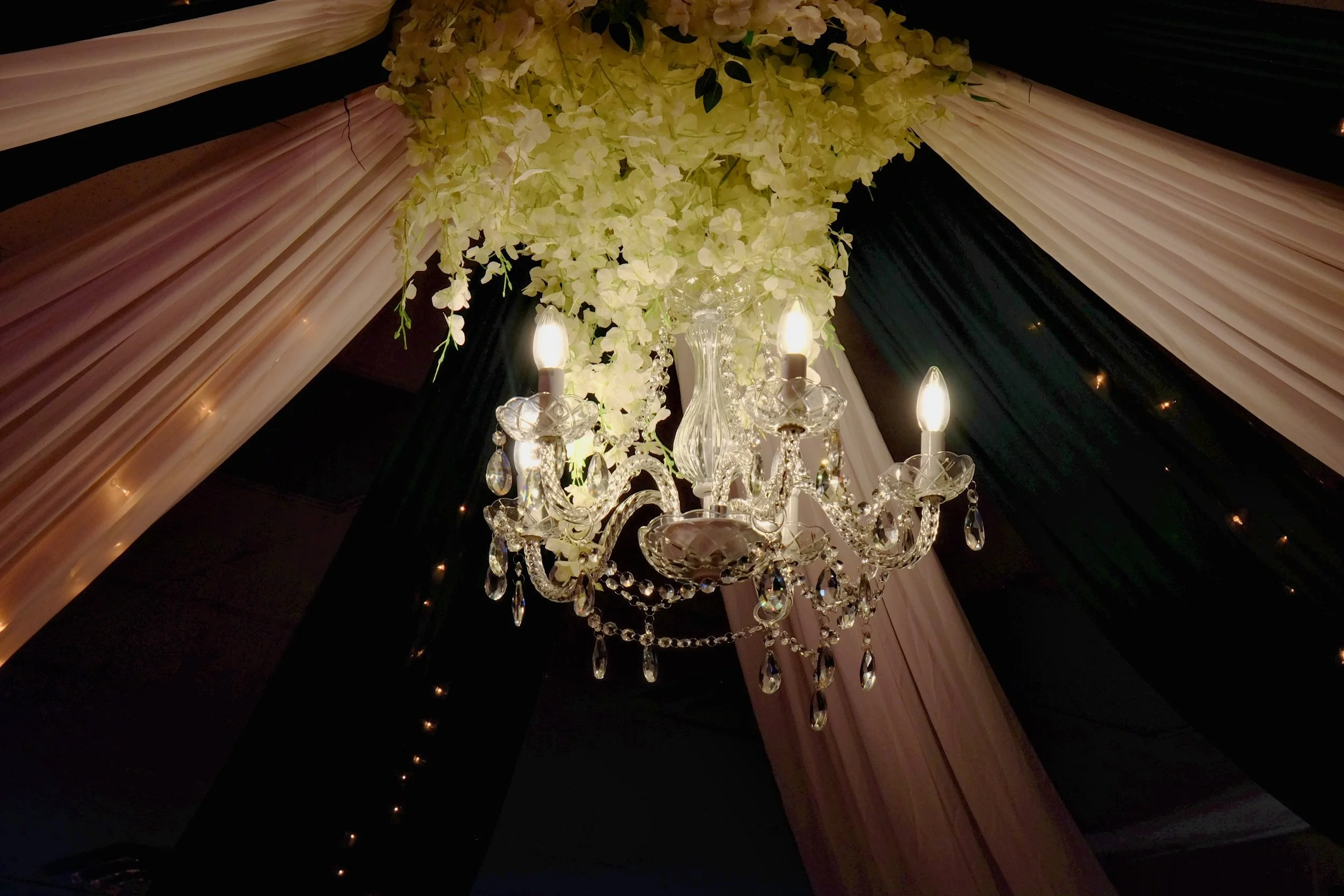 A chandelier decorated with white flowers and multiple crystal adornments, hanging from a ceiling draped with pink, black, and white fabric.