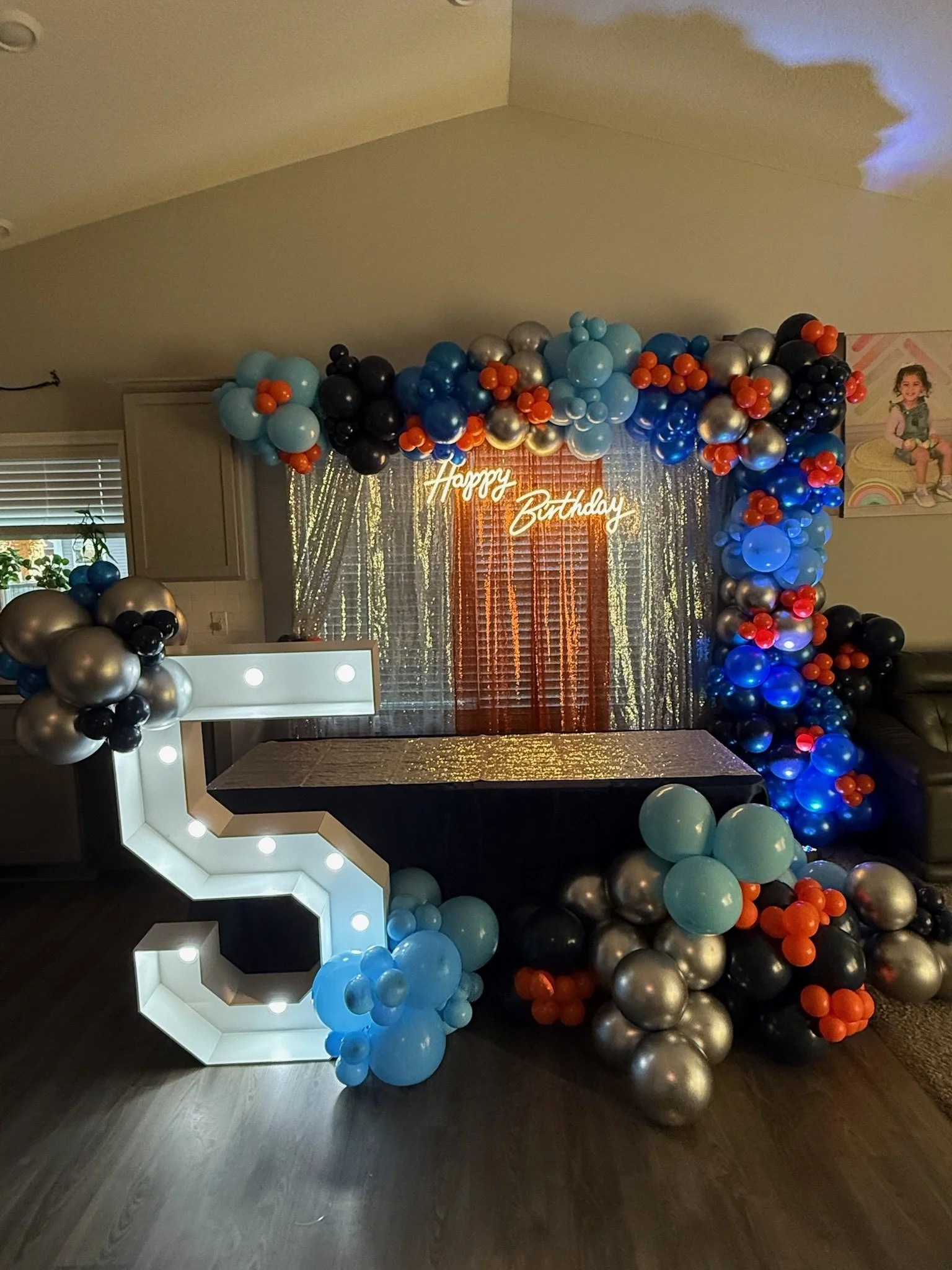 Colorful balloon arch frame with blue, black, silver, and orange balloons at a birthday celebration, with a neon 'Happy Birthday' sign in the background and a decorative backdrop.