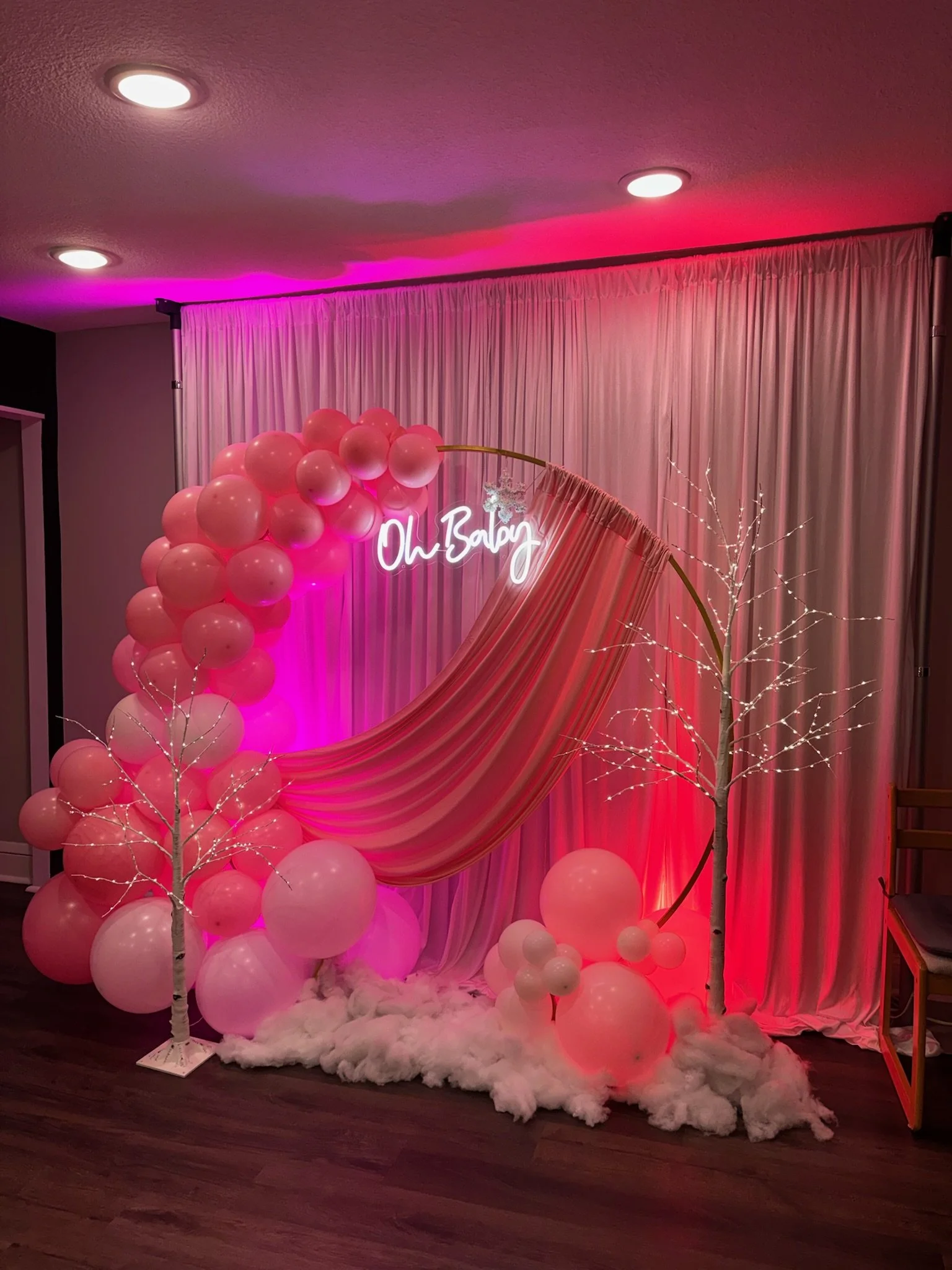 A decorative pink-themed setup with pink balloons, white artificial trees, pink drapes, pink neon sign that reads 'Oh Baby', and fluffy white material on the floor.