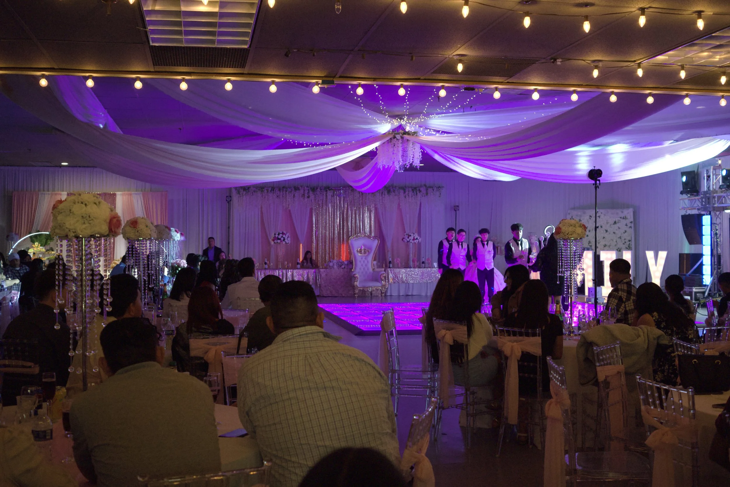 An indoor event hall decorated with purple and white drapery on the ceiling, illuminated by string lights and a chandelier. There are round tables with floral centerpieces, surrounded by guests. A stage with a decorated backdrop is at the front, with