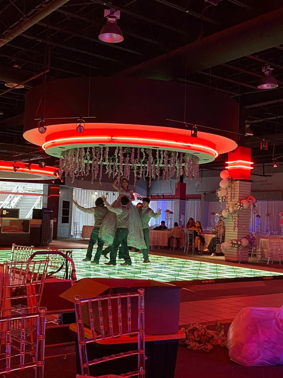 Group of children dancing on a lit-up dance floor at a celebration or party, decorated with hanging flowers and illuminated red ceiling fixtures, in a decorated event space with tables and balloons.