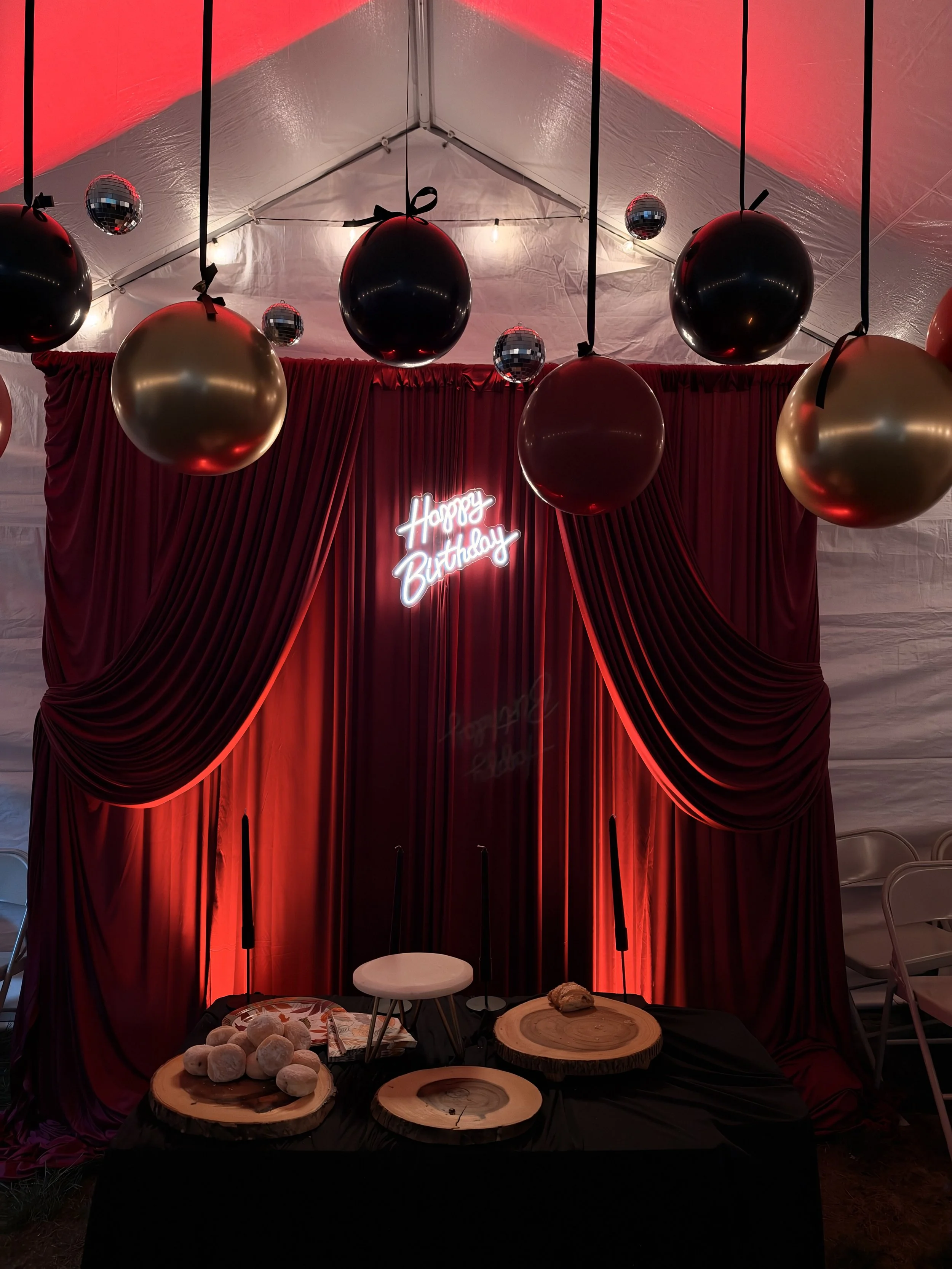 Decorative table setting with a red curtain backdrop and a neon sign that says "Happy Birthday." Balloons, including black, gold, and disco ball balloons, are hanging from the ceiling. On the table, there are wooden platters with snacks, pinecones, and a white chair is visible on the right side.