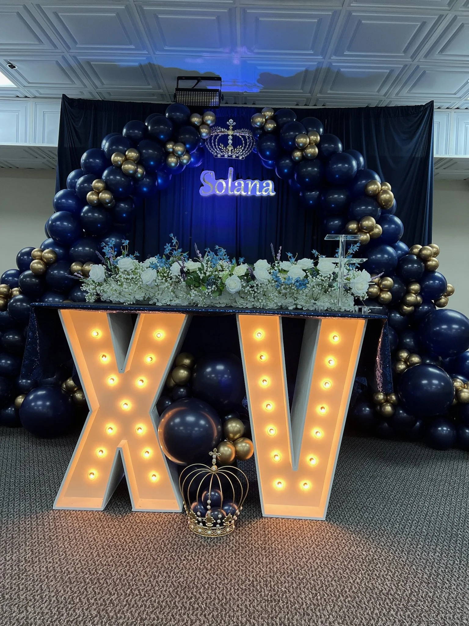 Decorative setup with blue and gold balloons, a floral arrangement, a large illuminated letter V, a crown, and a sign with the name Solana at an event venue.