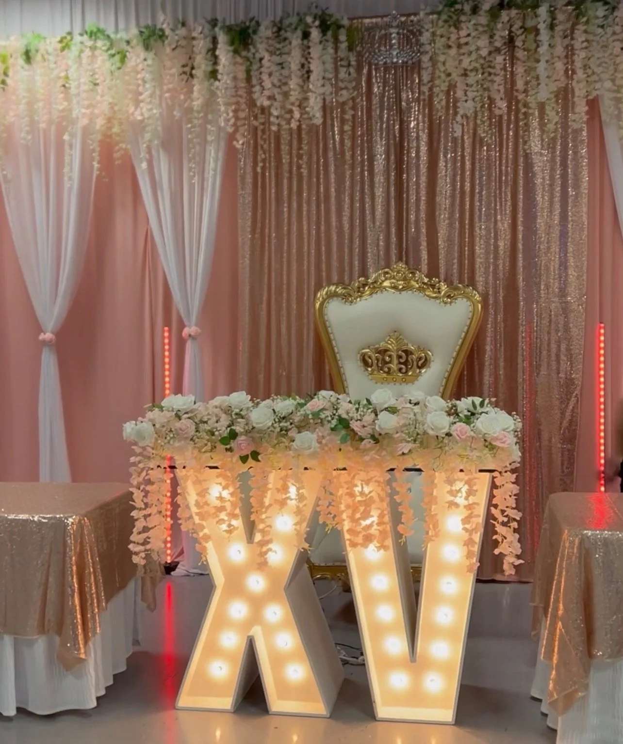 Elegant bridal or event setup with a white and gold throne-like chair behind a table decorated with white and pink flowers, illuminated marquee letter W in front, pink and gold draped backdrop, and floral arrangements.