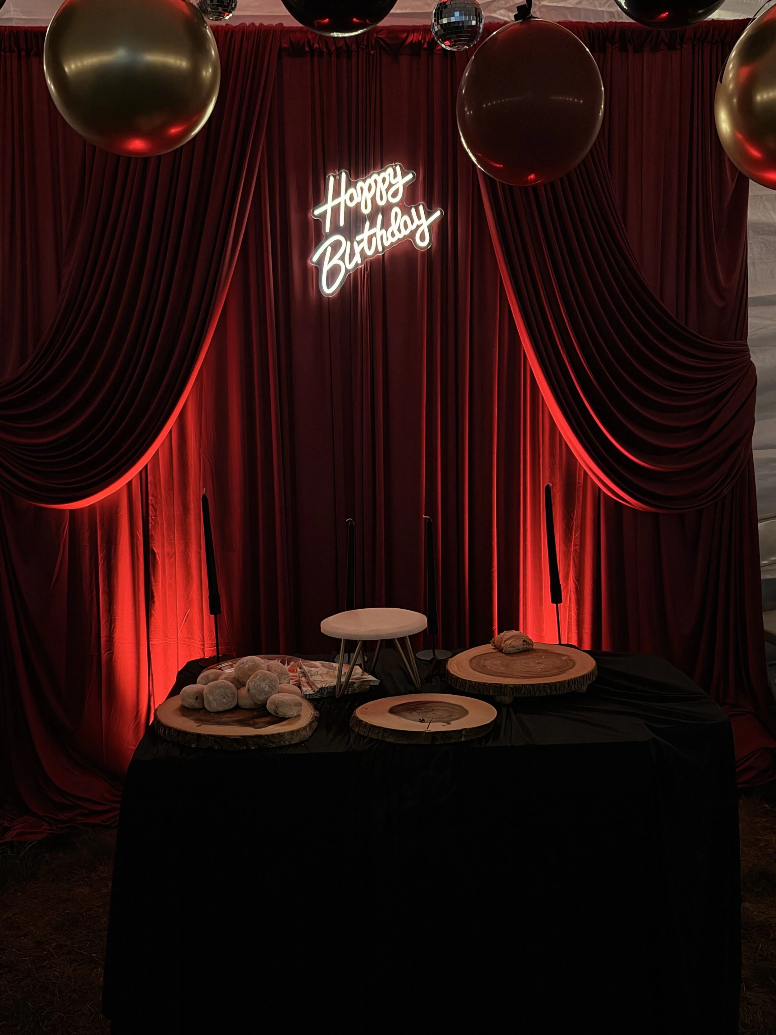 Decorative backdrop with red curtains and a neon sign that says 'Happy Birthday,' with large black and gold balloons suspended overhead. There are wooden slabs on a table with some gray rocks or stones on top, creating a rustic birthday celebration s