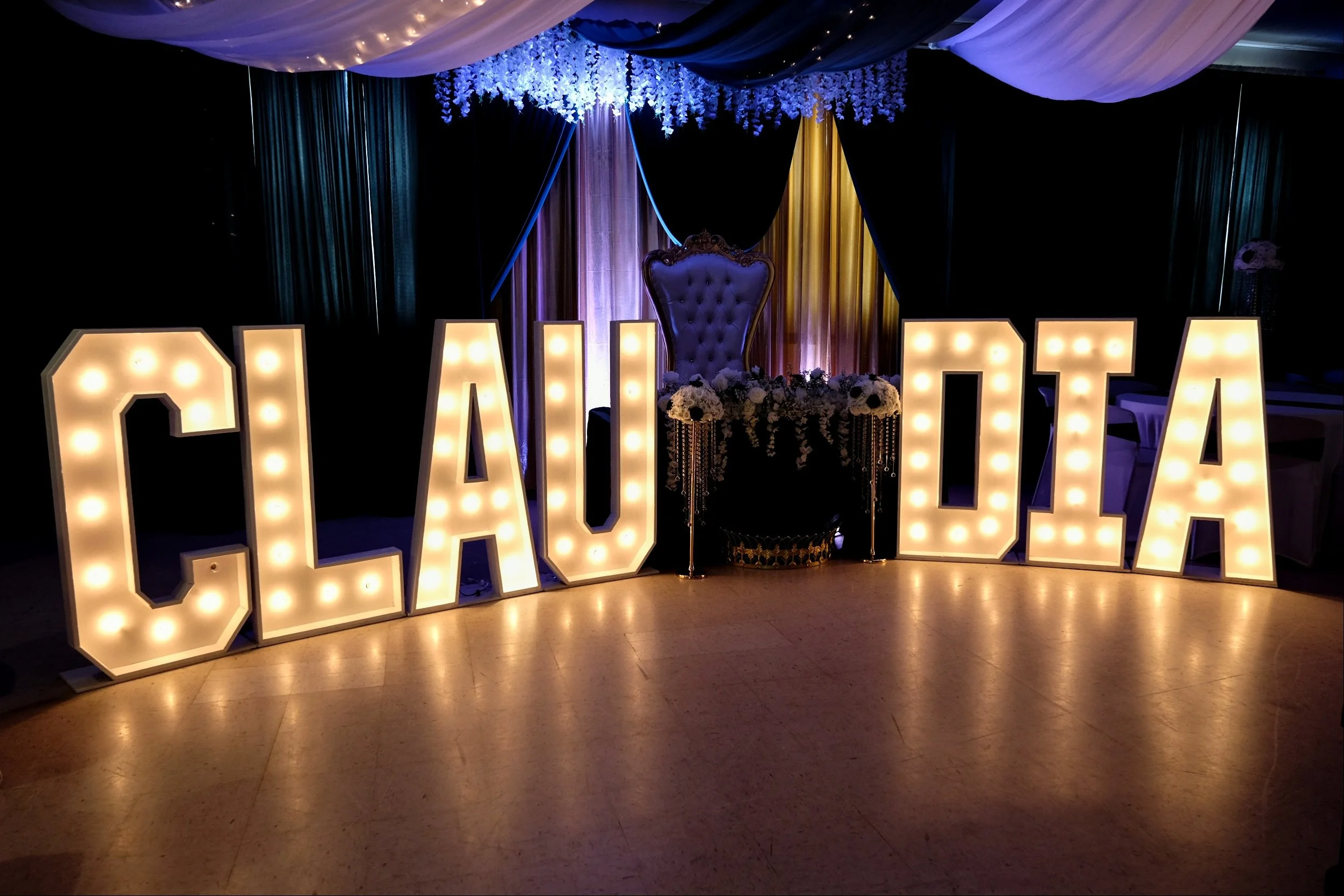 Large illuminated marquee letters spelling 'GAMER', with a decorated stage background featuring floral arrangements, a velvet throne, and draped fabric in purple, black, and gold colors.