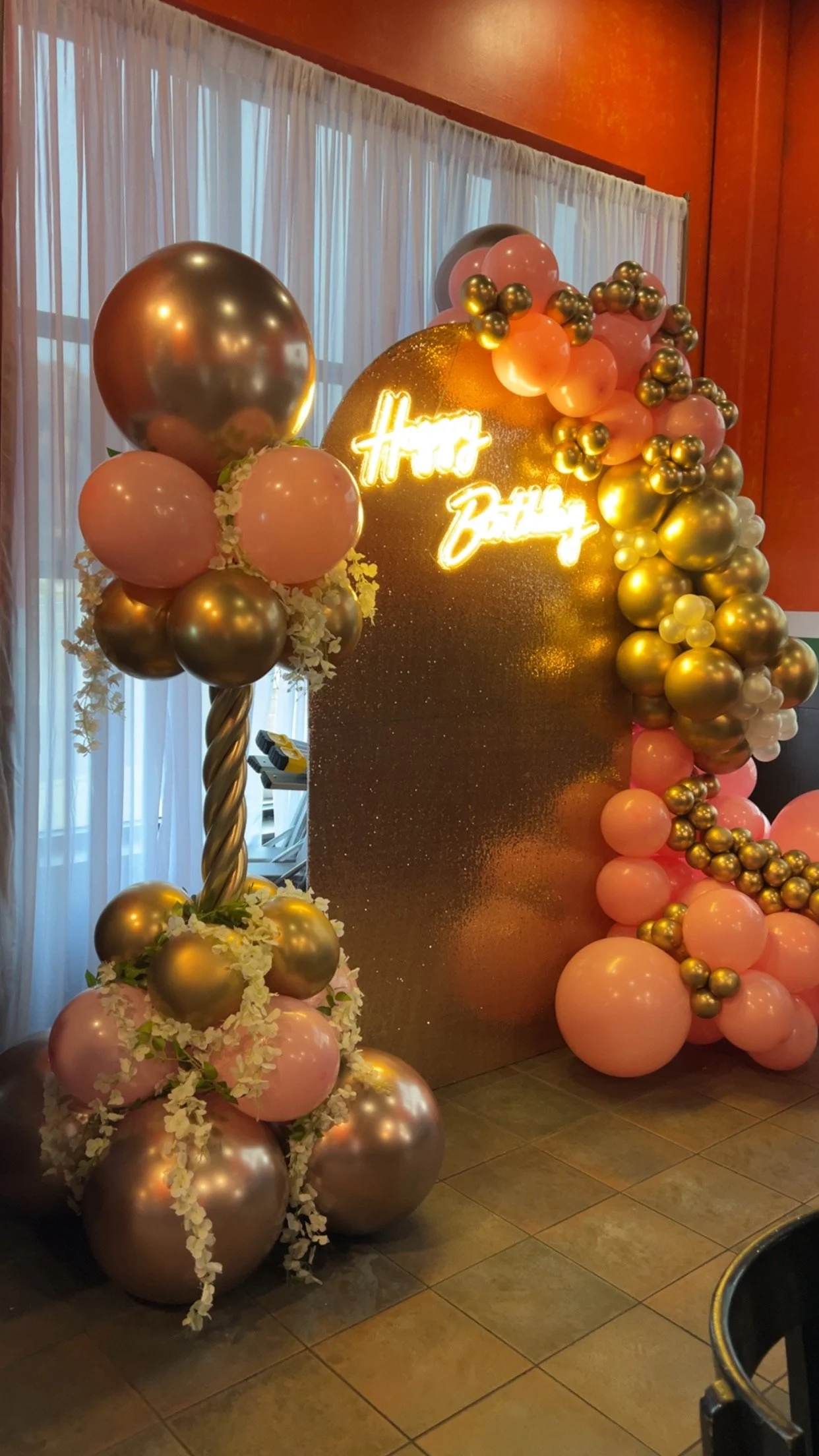 Decorative birthday display with pink, gold, and silver balloons arranged around a glittery backdrop that has a neon sign reading 'happy birthday.' A balloon column with pink and gold balloons topped with a twisted balloon design is also visible.